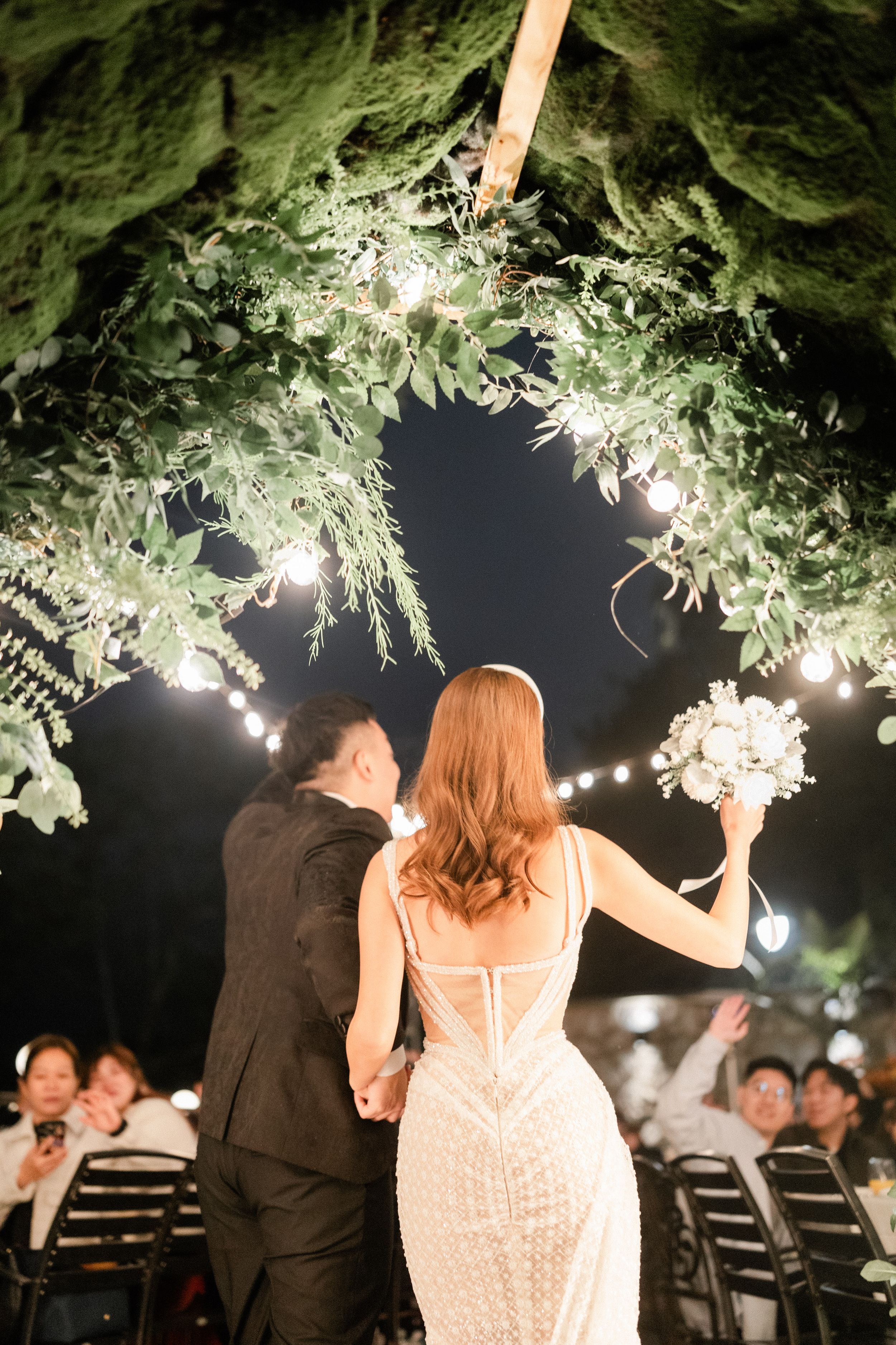 Bride and groom holding hands under floral arch with string lights at outdoor wedding reception.