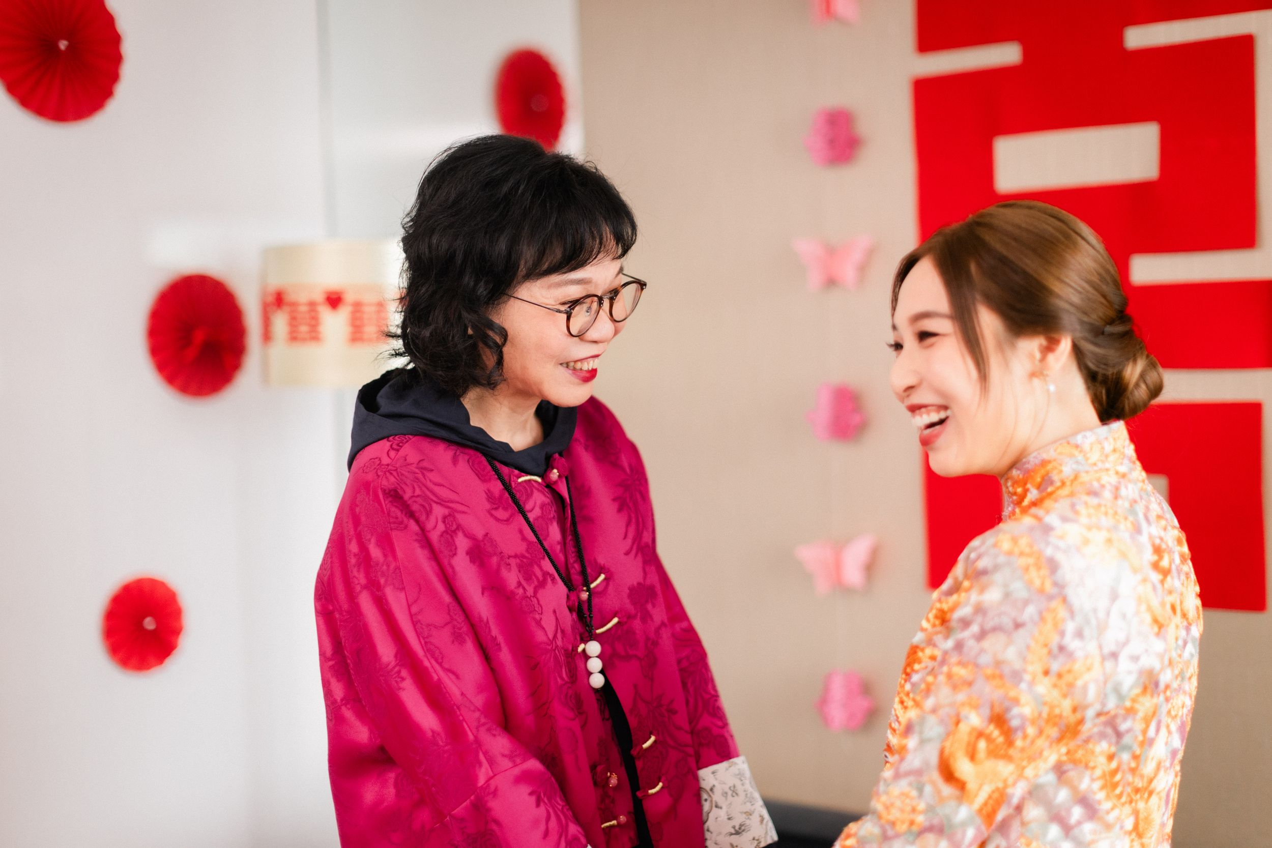 A smiling bride in qipao shares a joyful moment with an older woman in pink at a Chinese wedding.