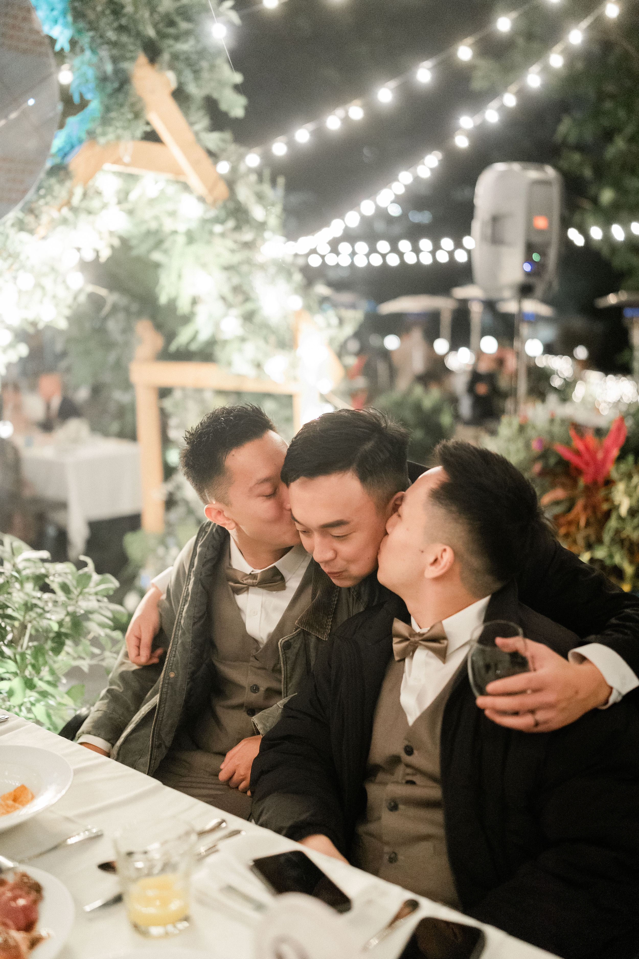 Three men in suits sharing kisses at a wedding reception dinner table with string lights.