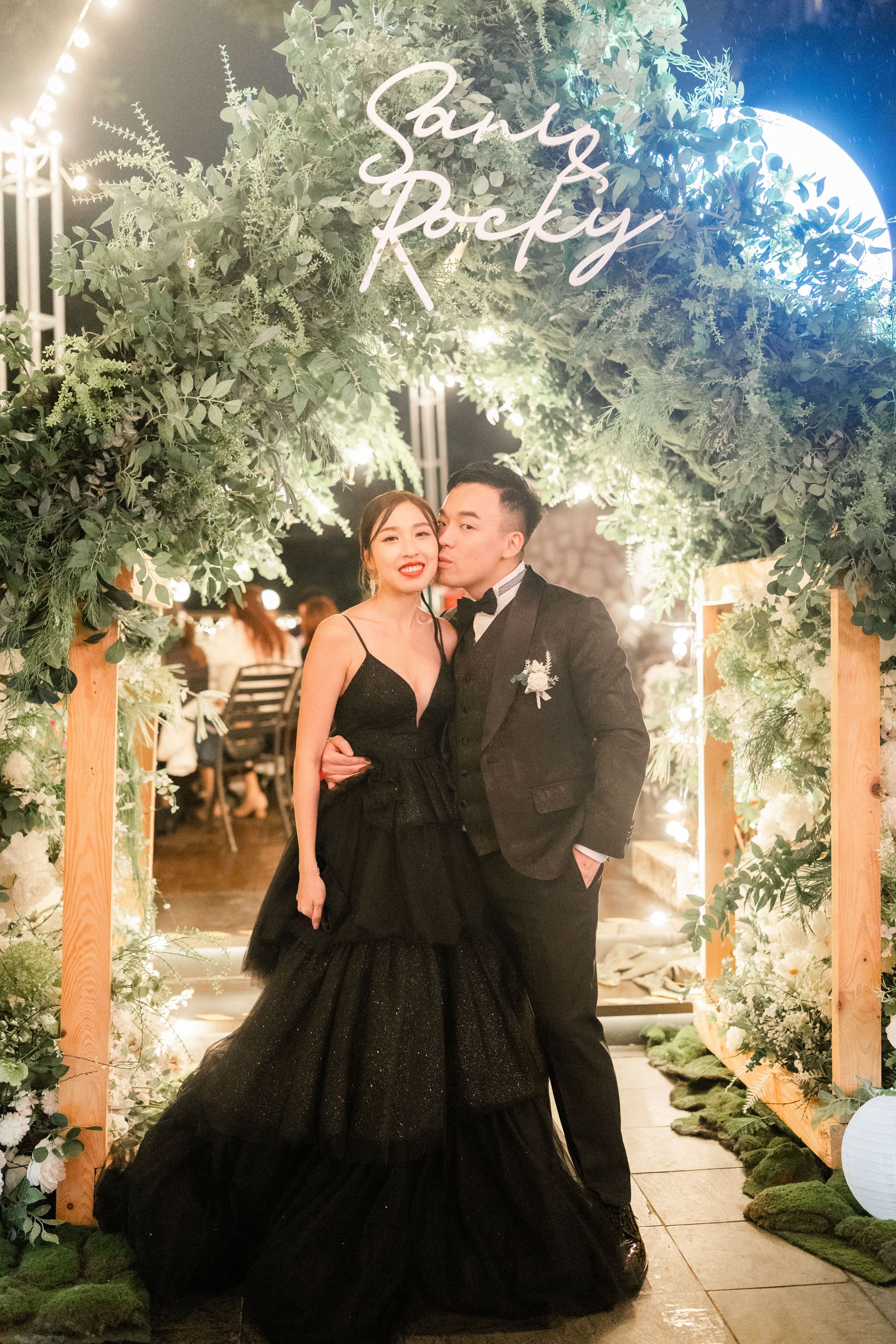 Couple posing under neon sign reading San Rocky at elegant outdoor wedding reception at night.