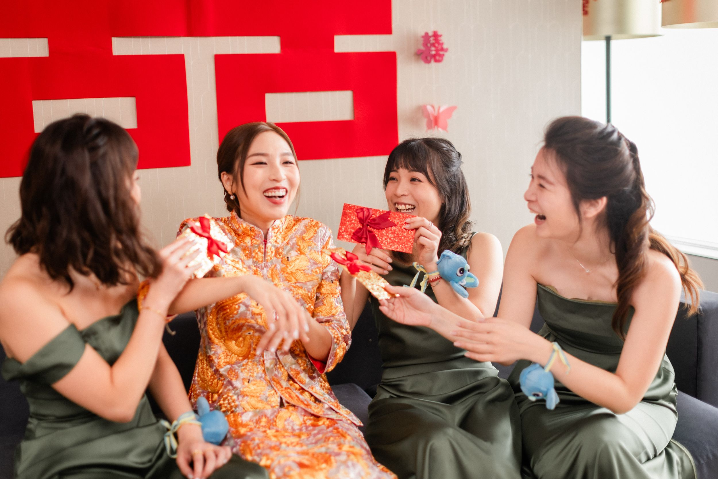 A bride in orange qipao laughs with bridesmaids in olive green as they exchange red envelopes at a Chinese wedding.