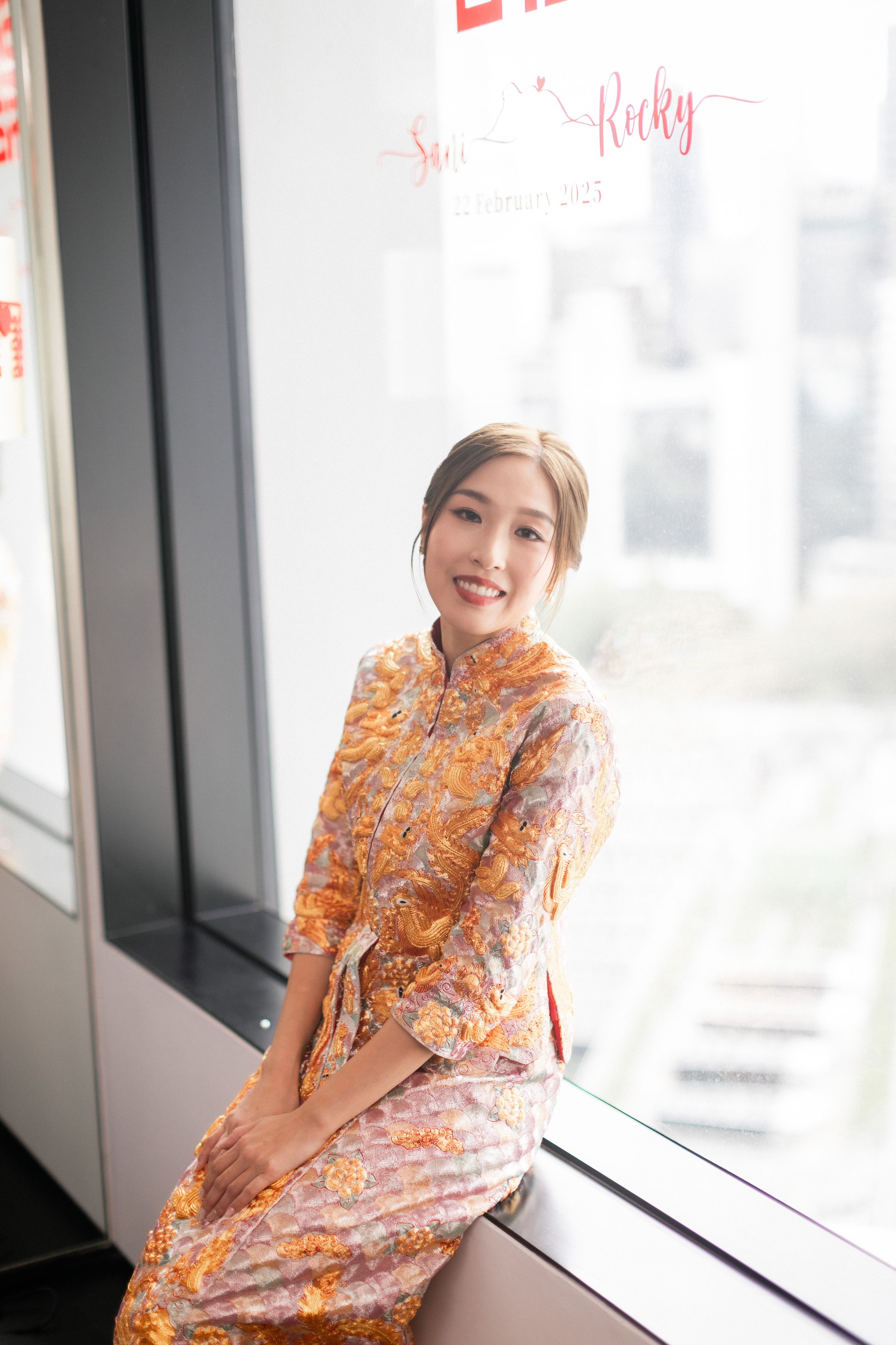 A young woman in a floral orange qipao sits by a window, smiling gracefully at a Chinese wedding.