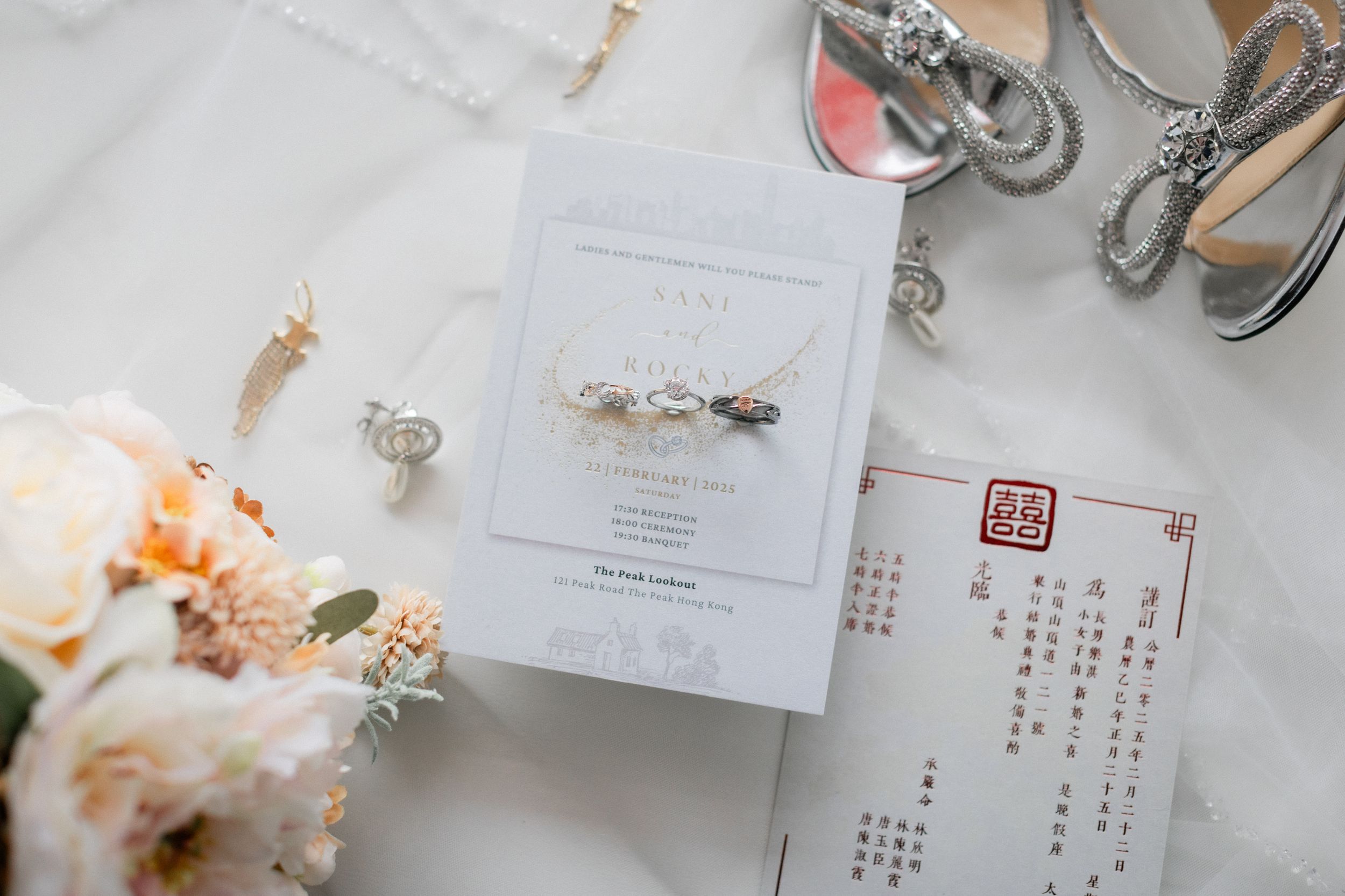 Wedding flat lay with white invitation, pearl necklace, floral bouquet, and Chinese ceremony card.