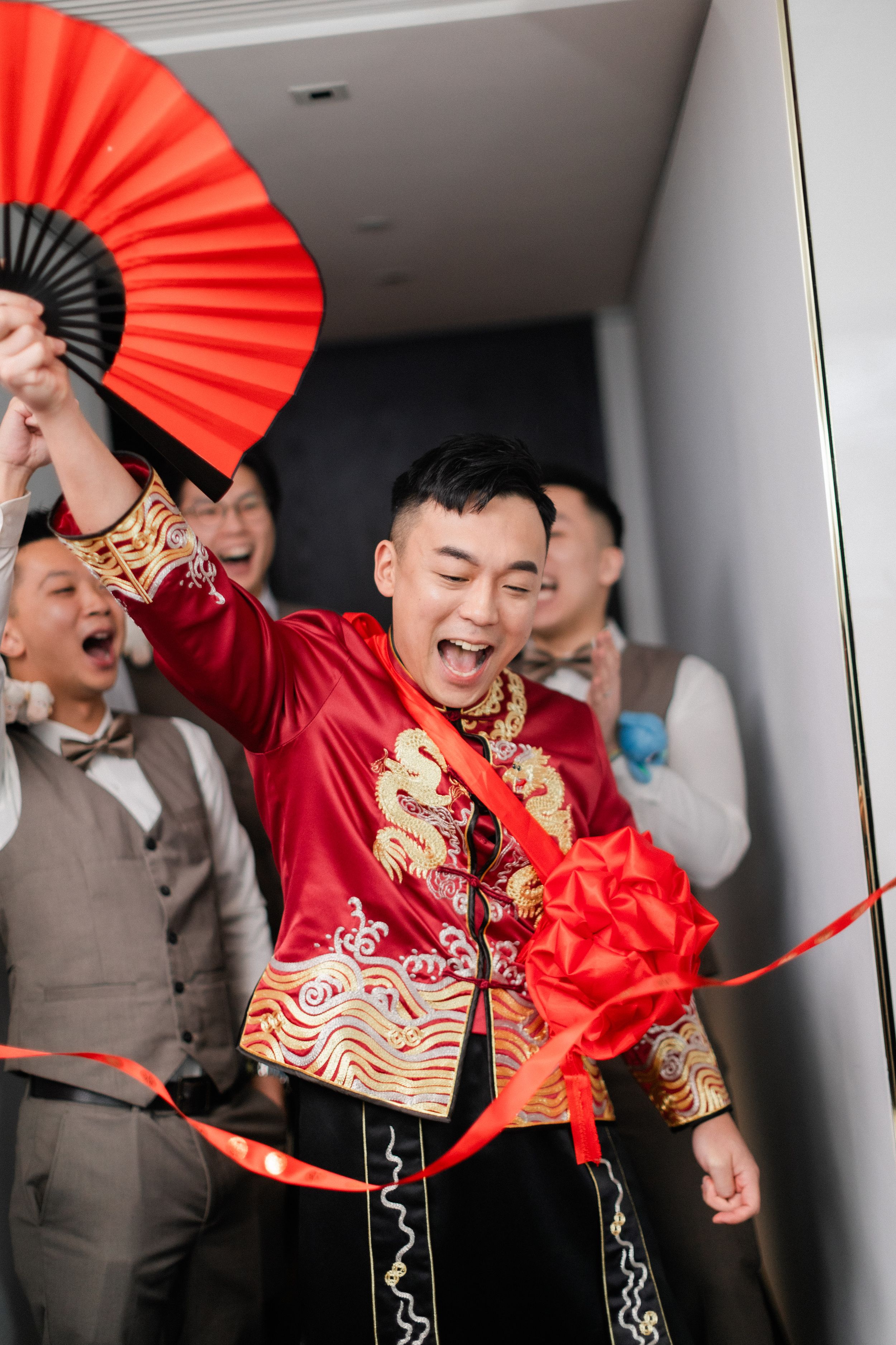 An excited groom in red dragon embroidered qipao waves a red fan while breaking through a ribbon at his wedding.