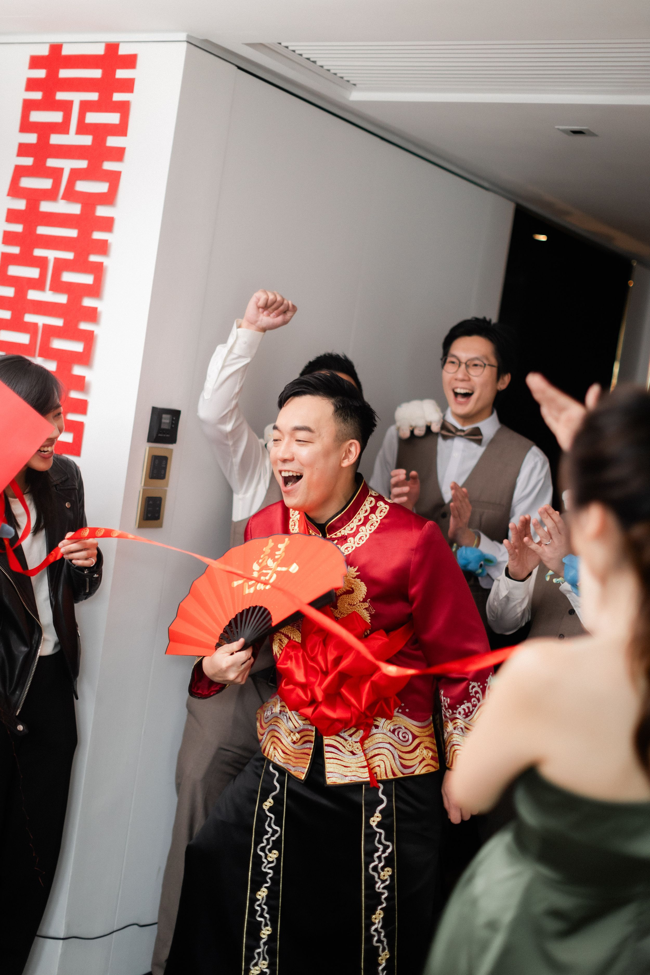 A jubilant groom in traditional red Chinese wedding attire waves a fan as groomsmen celebrate behind him.
