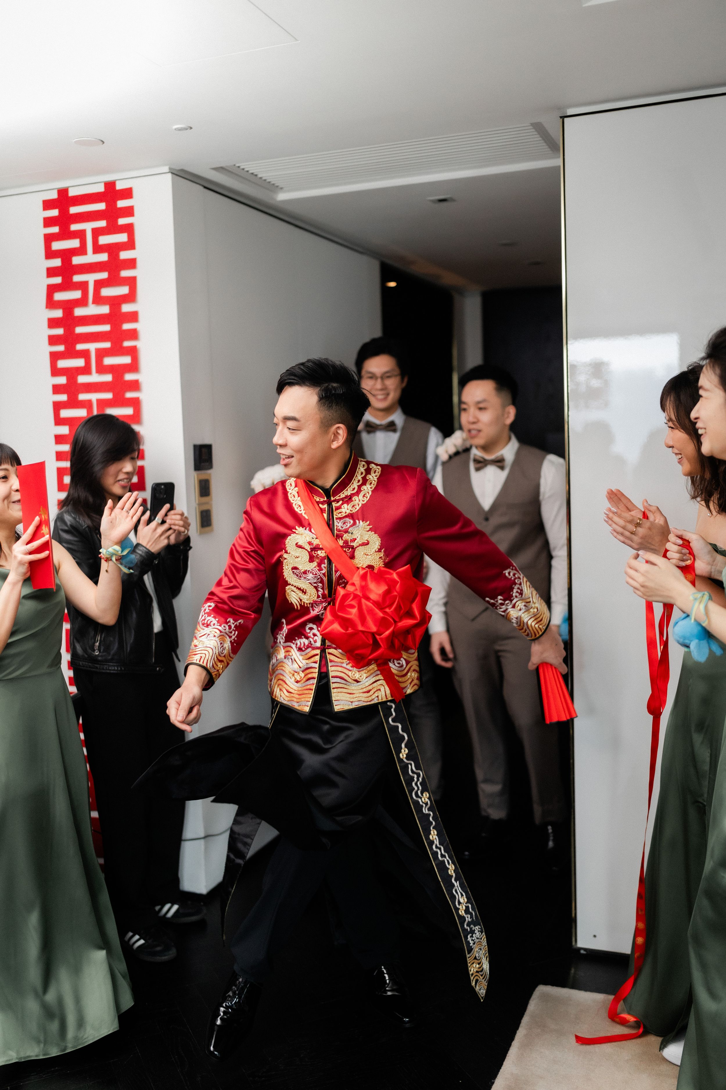 A groom in a red embroidered Chinese wedding suit makes a grand entrance as guests cheer and applaud.