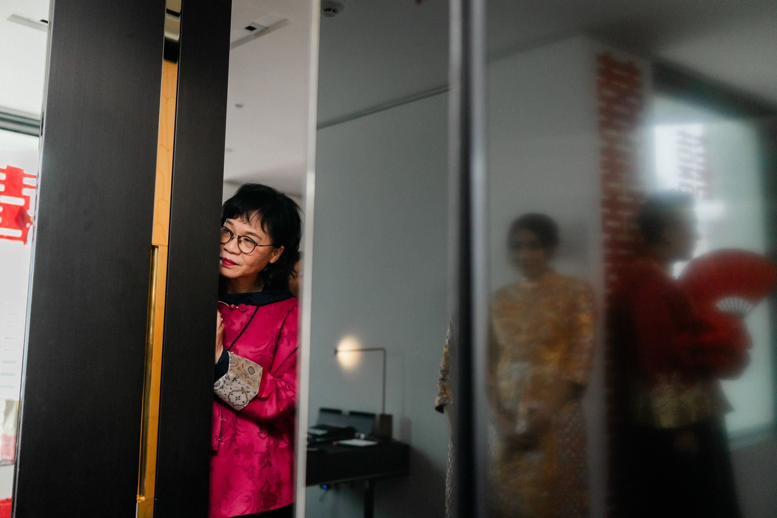 Woman in pink traditional Chinese dress peeks through a doorway during a wedding ceremony.
