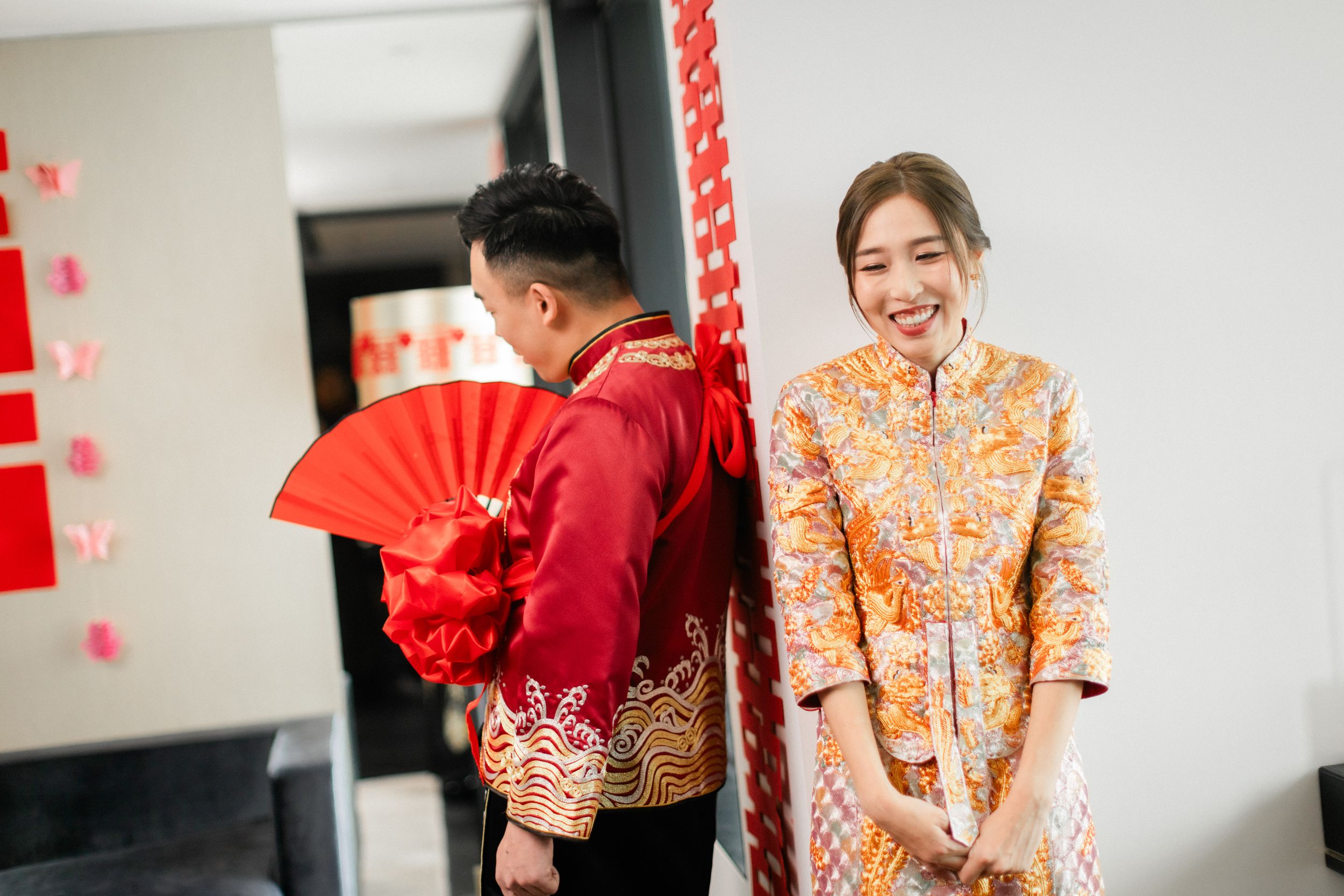 Smiling bride in gold qipao stands beside groom holding red fan during Chinese wedding.