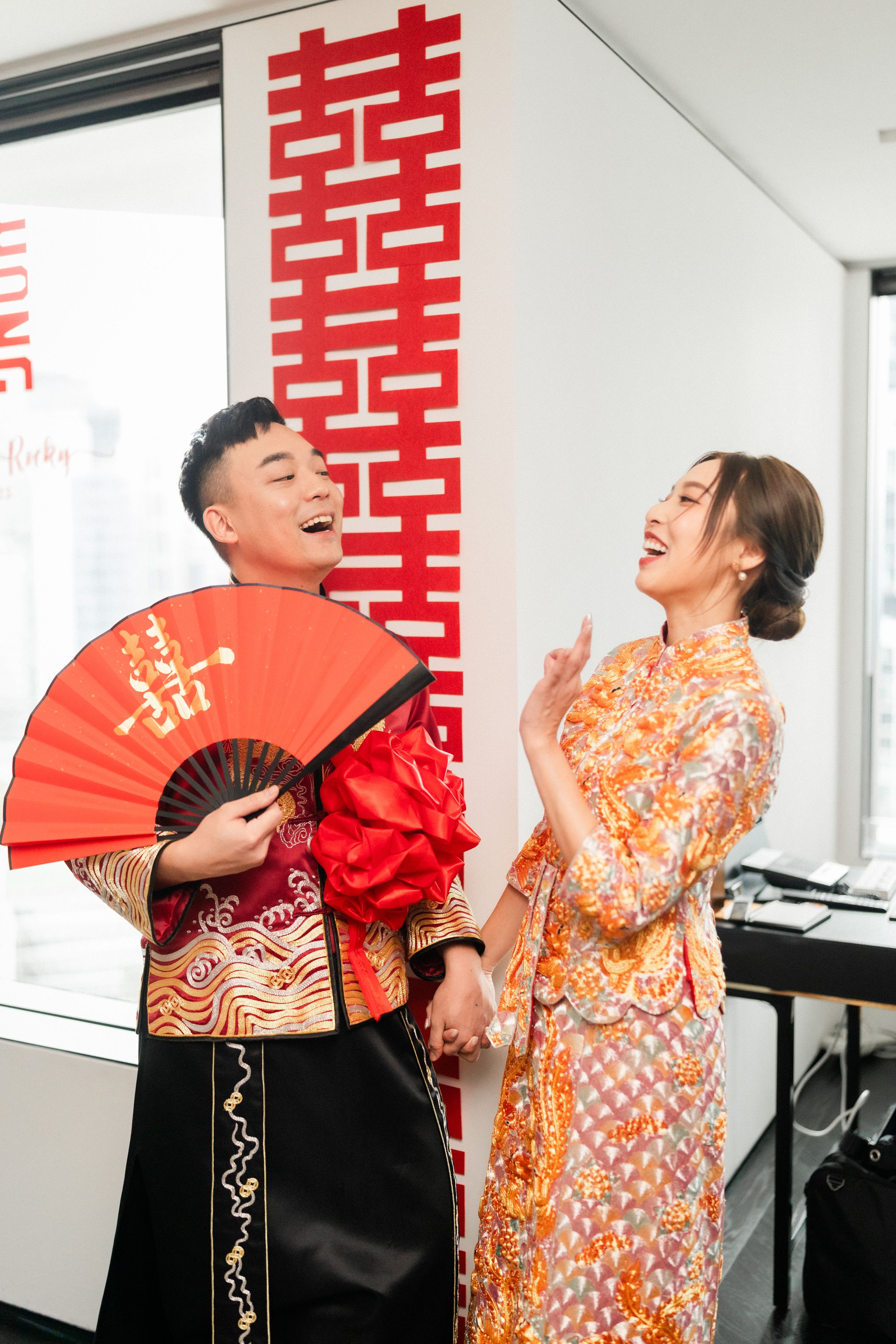 Laughing Chinese wedding couple in traditional attire with red fan beside double happiness symbol.