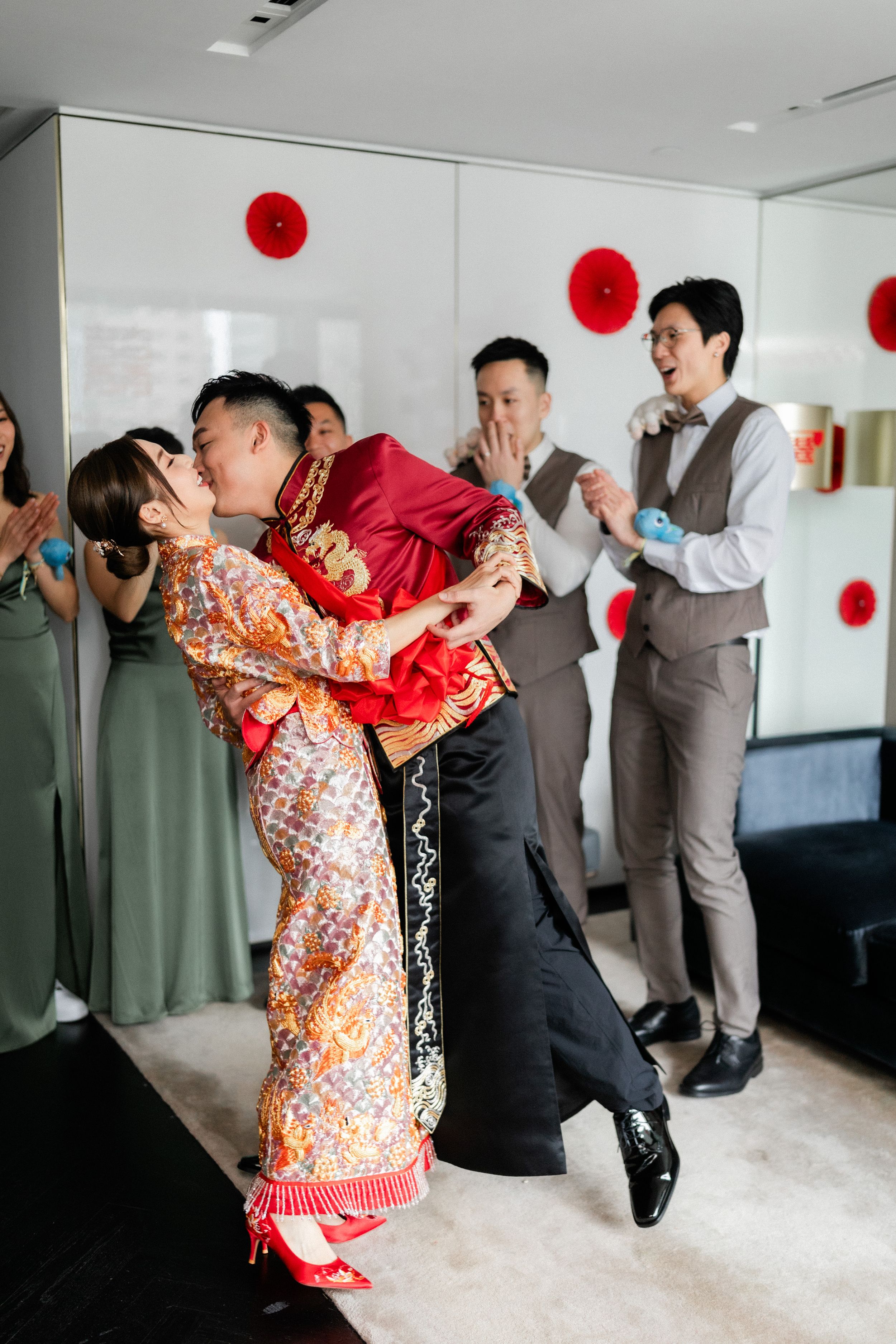 Groom lifts bride in traditional Chinese wedding attire as guests cheer and applaud.