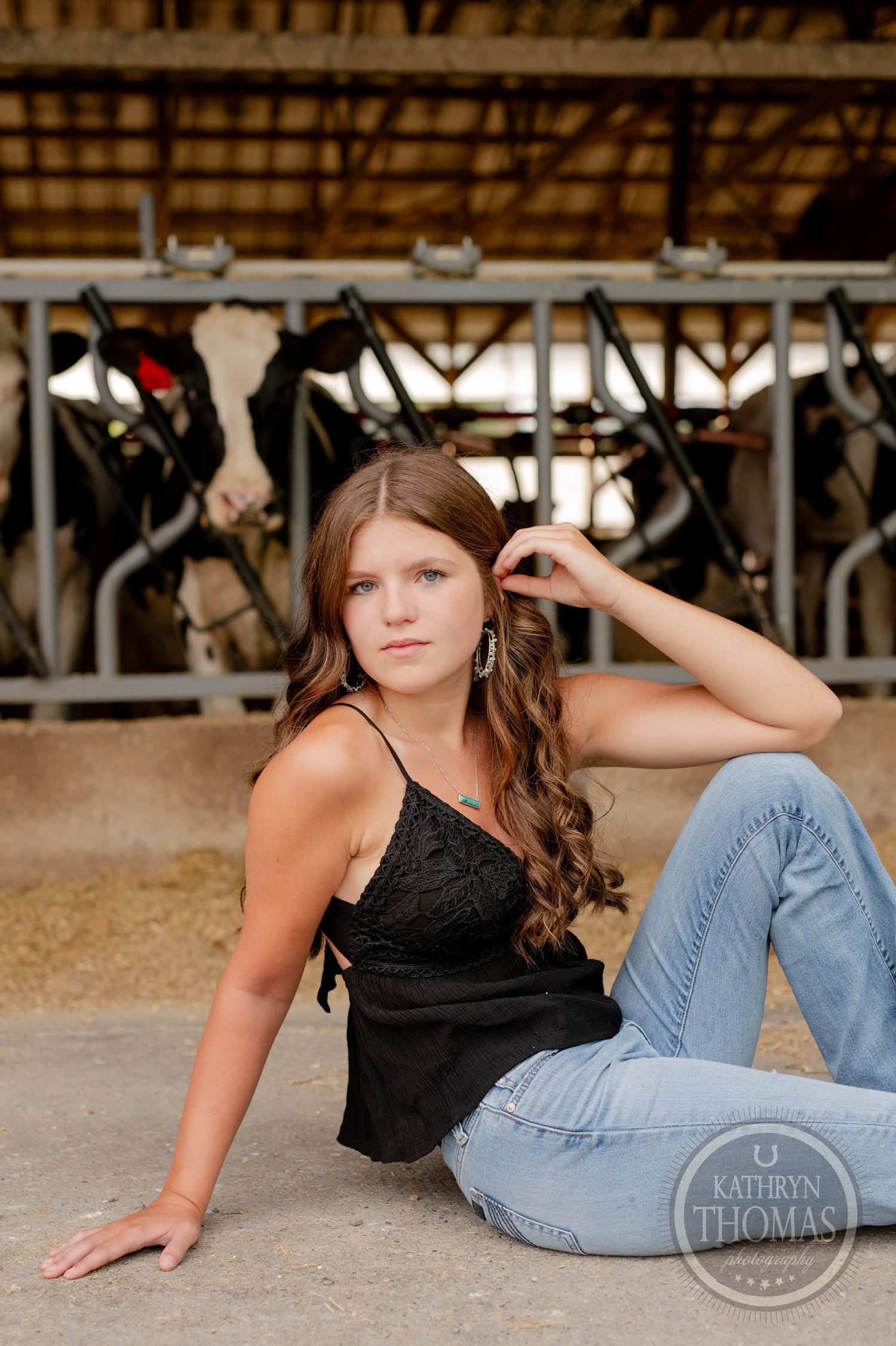Relaxed pose on barn floor wearing black lace top with dairy cows visible behind.
