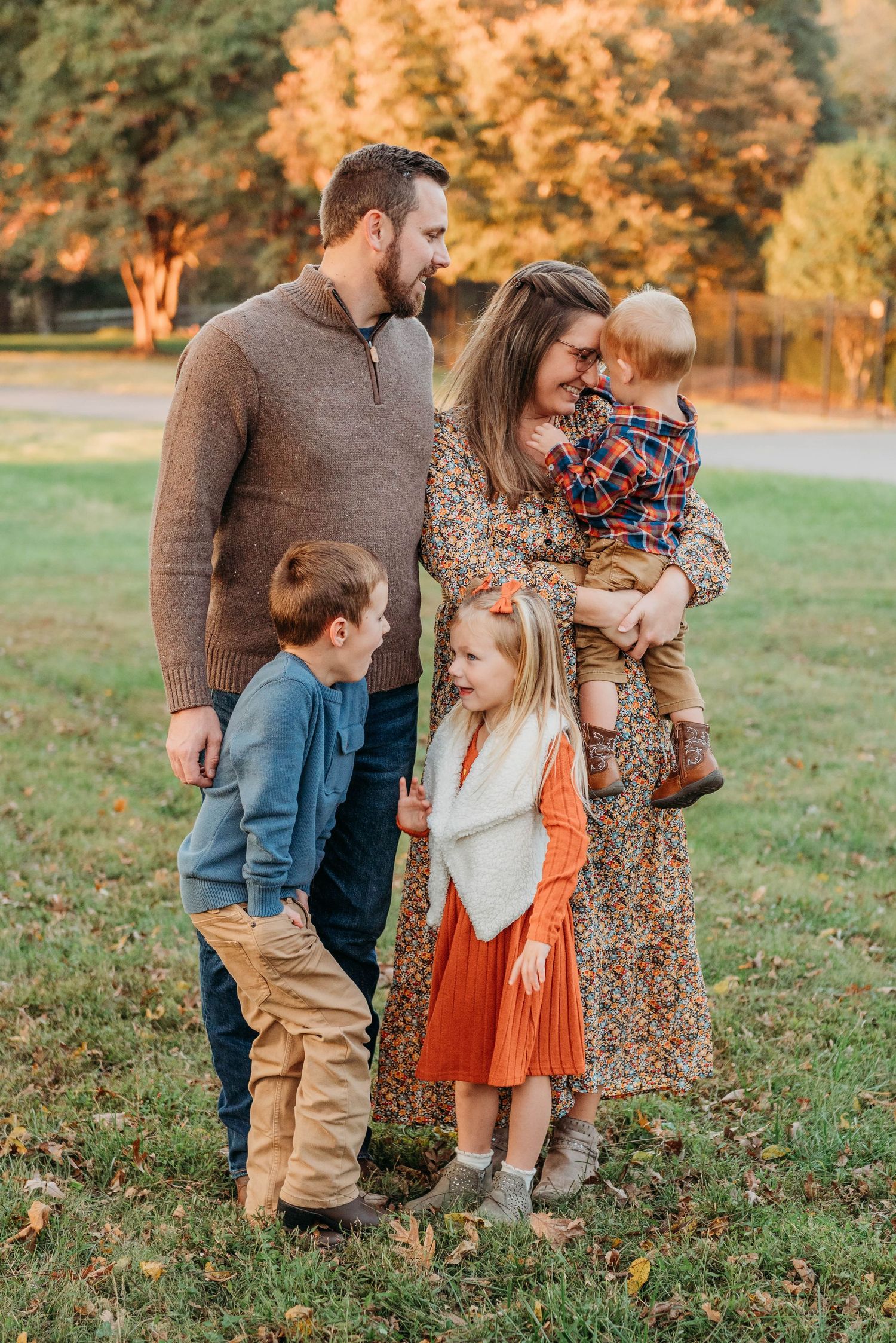 A group wearing coordinating fall outfits stands together outdoors with autumn foliage in the background during sunset.