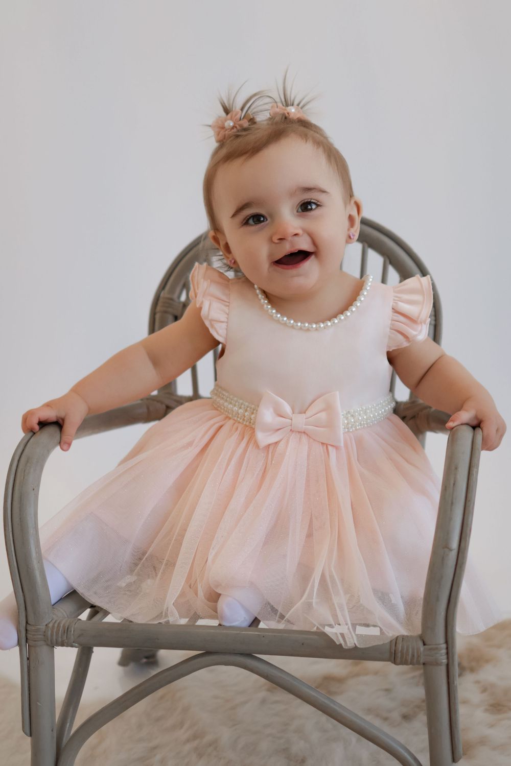 Baby in peach tulle dress with bow detail sitting on gray chair.