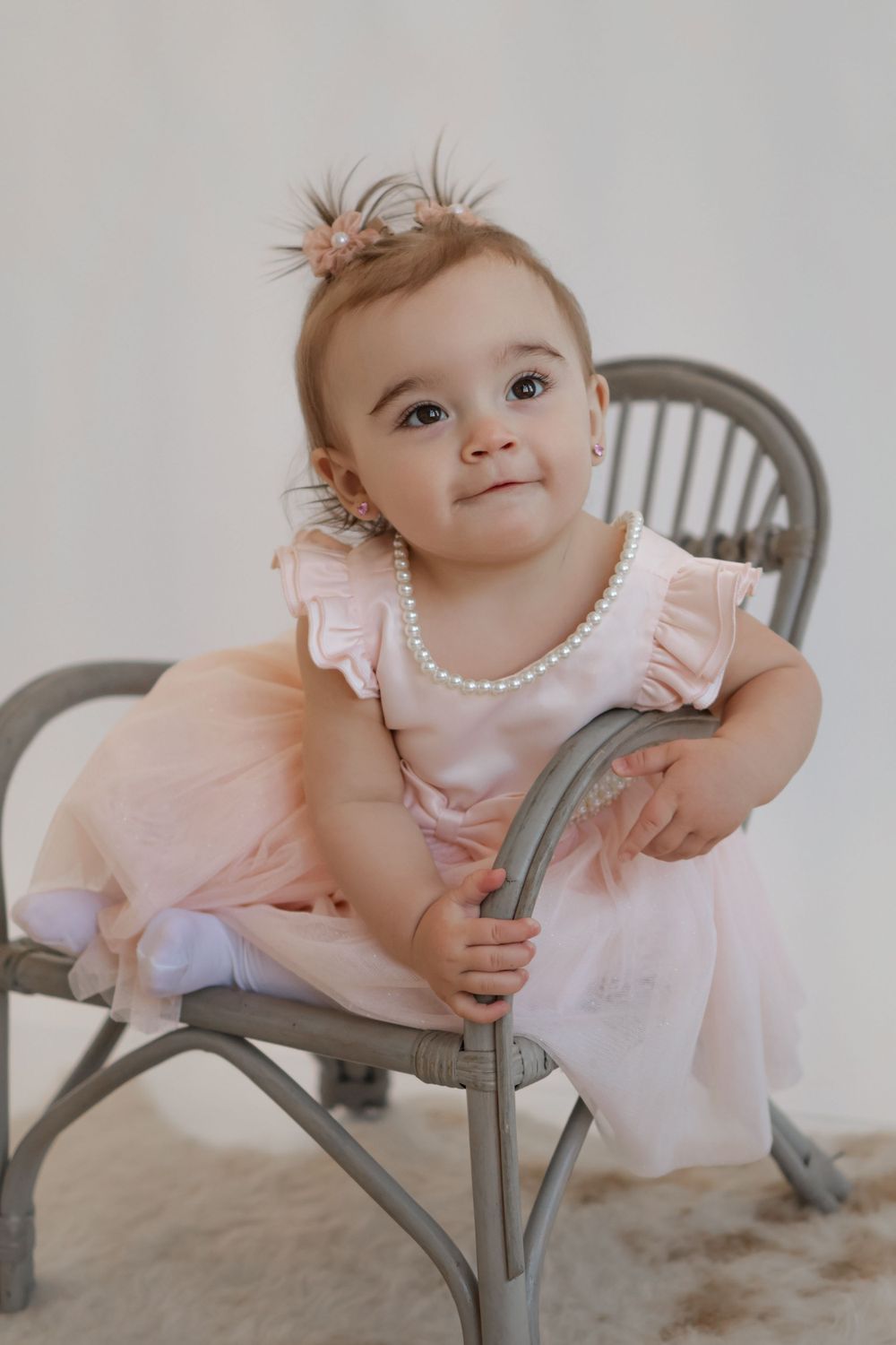 Baby in pink dress with ruffled sleeves posing on gray chair.