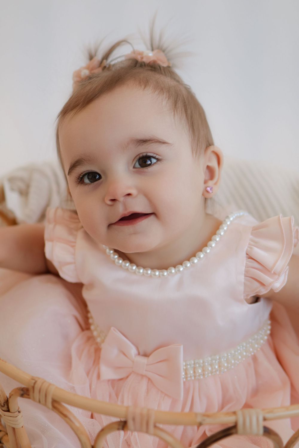 Baby in pink dress with bow detail and pearl necklace sitting in wicker chair.