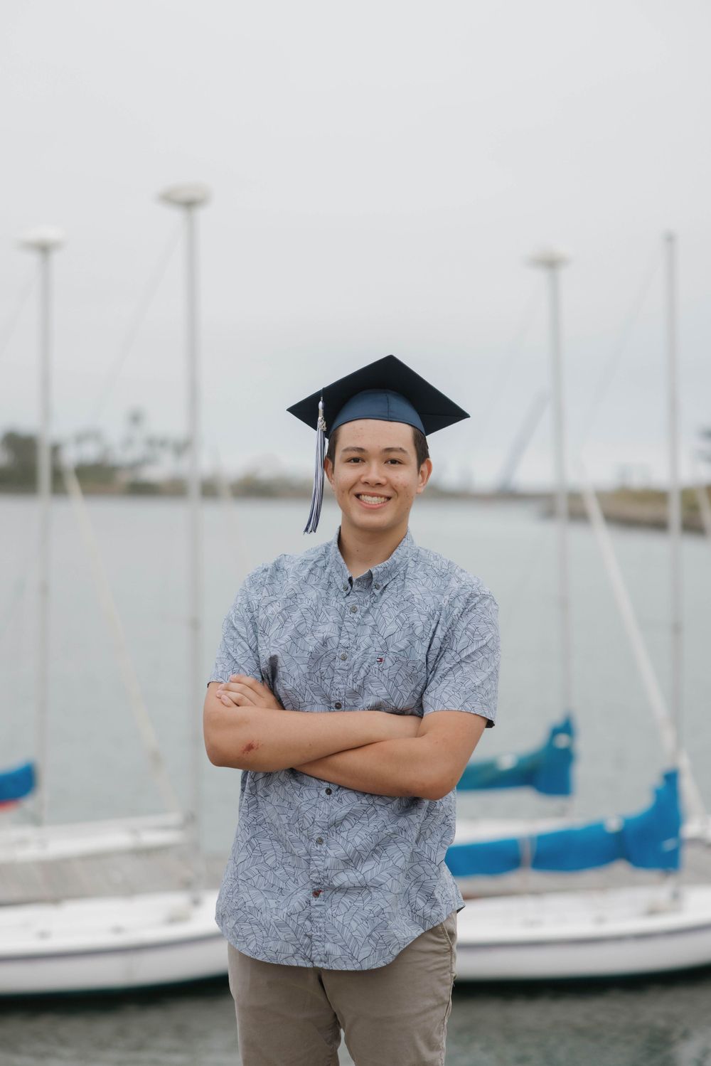 A graduate wearing a mortarboard cap and gray shirt stands at a marina with sailboats in the background.