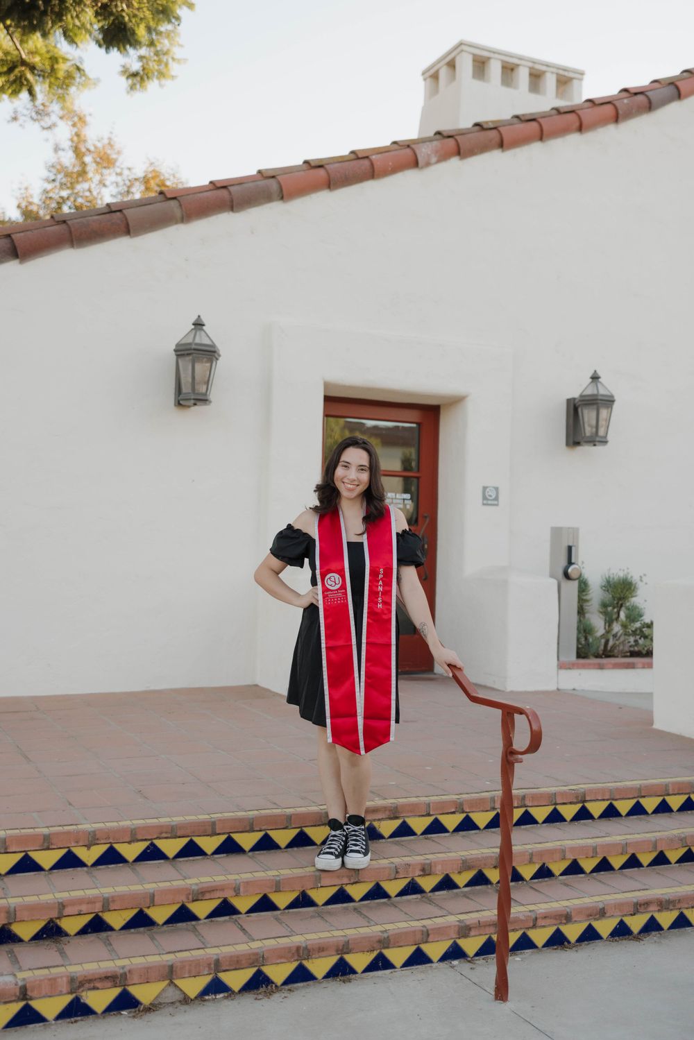 Graduate in black dress with red stole stands on decorative Spanish-style tiled steps outside white stucco building.