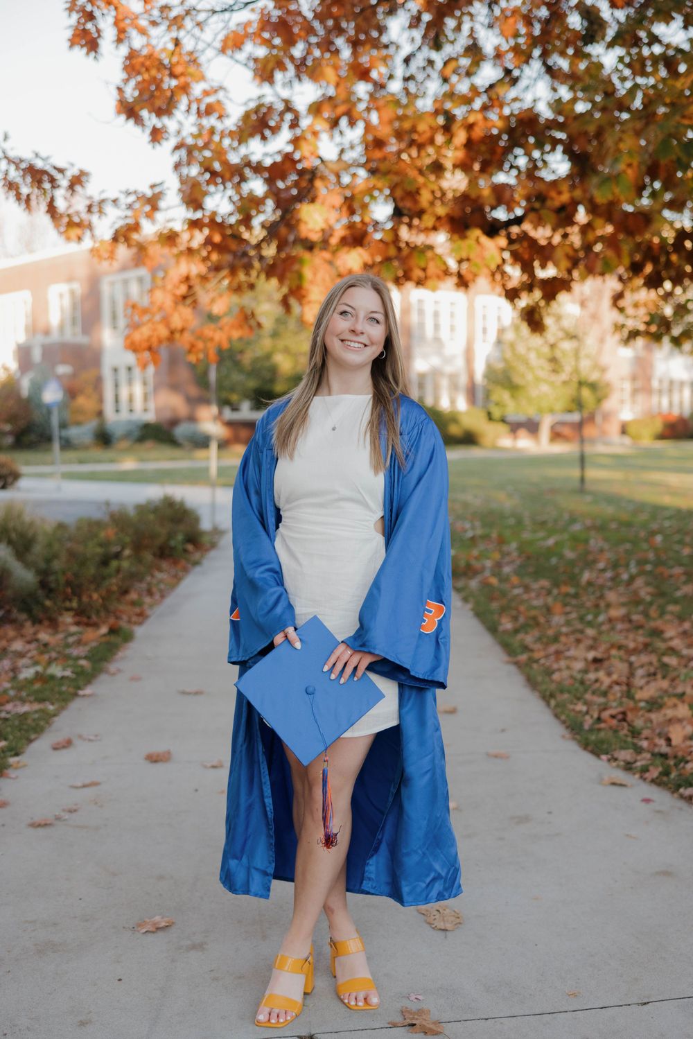 Graduate in blue cap and gown stands on campus sidewalk during autumn with orange foliage in background.