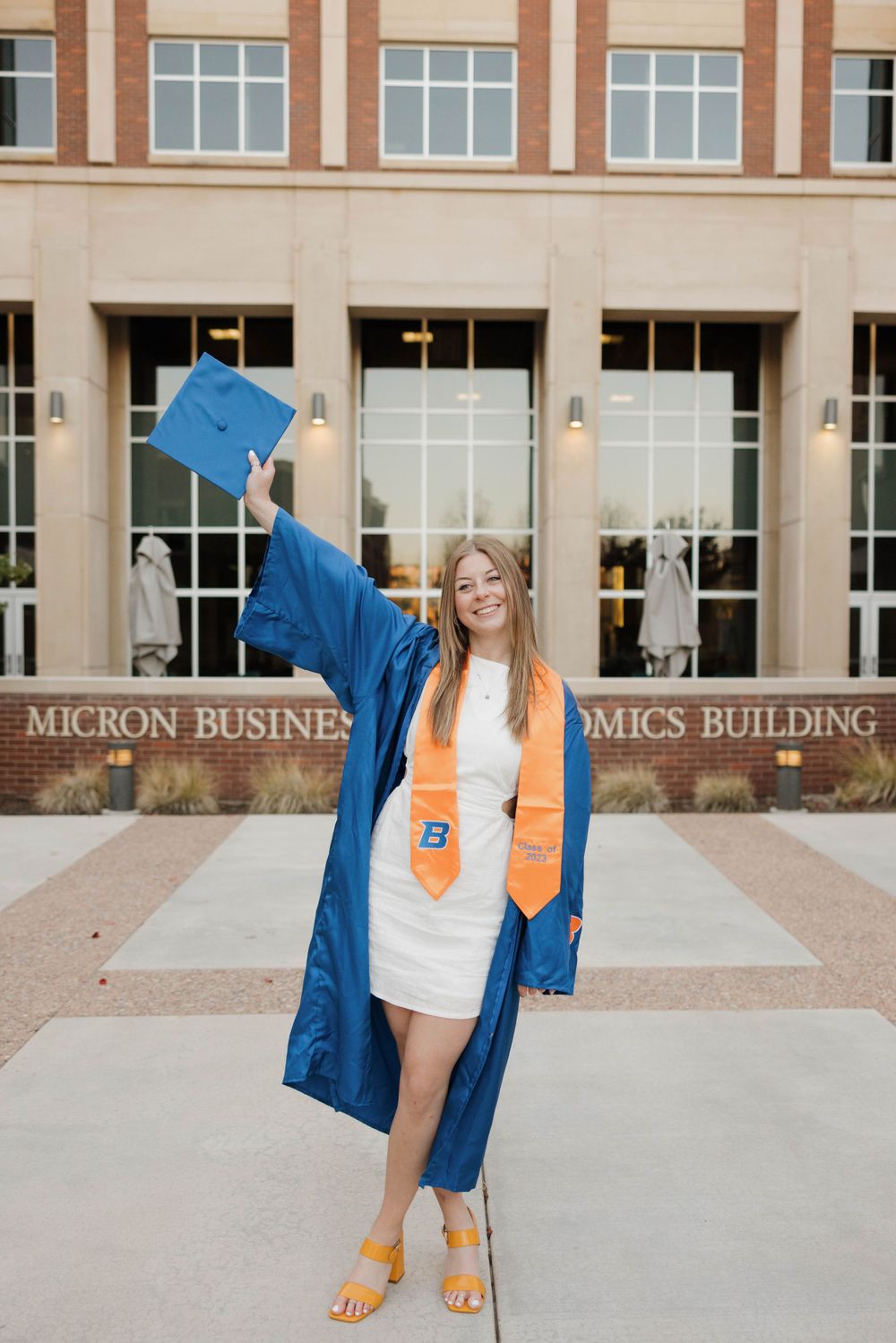 A graduate in blue cap and gown celebrates outside the Micron Business Economics Building.