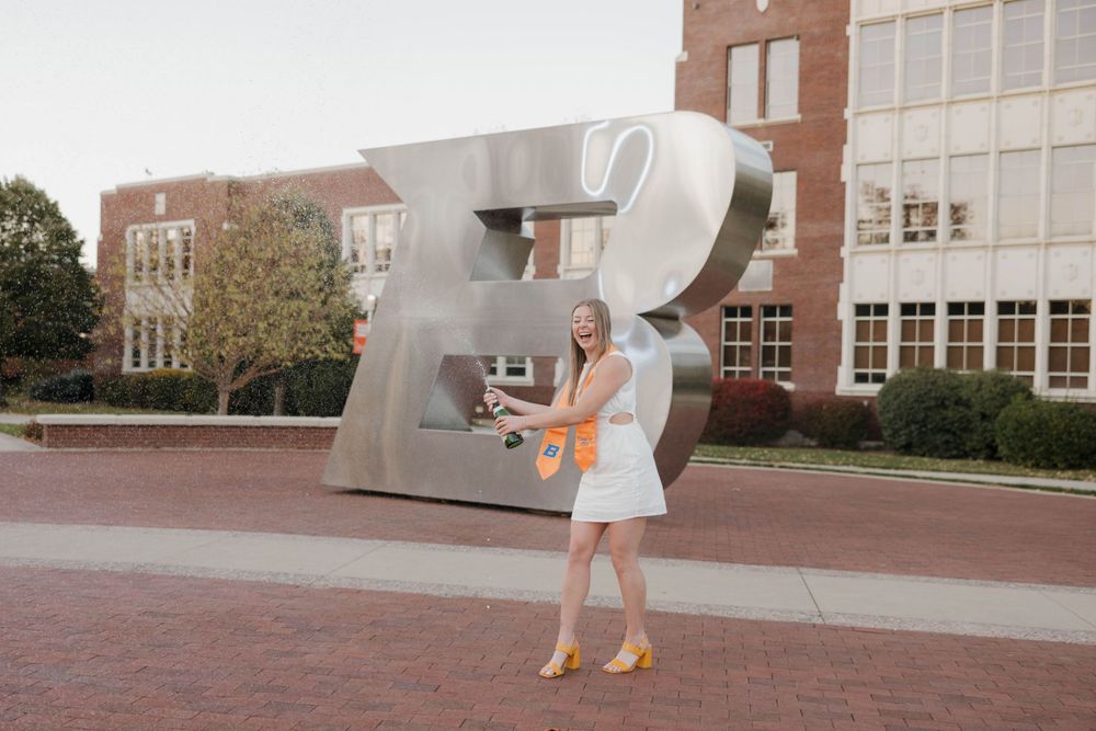 Someone poses with a large silver question mark sculpture outside a brick university building.