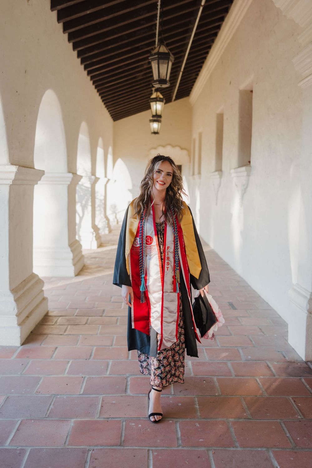 Graduate in traditional academic regalia with colorful stole walks through historic arched hallway with terracotta floors.