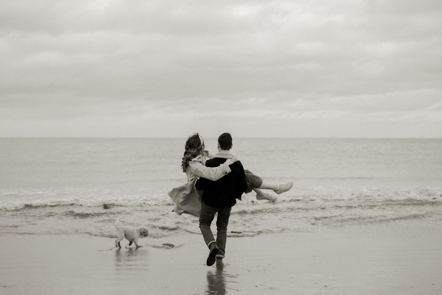 A romantic black and white beach scene as two people dance by the shore with a dog nearby.