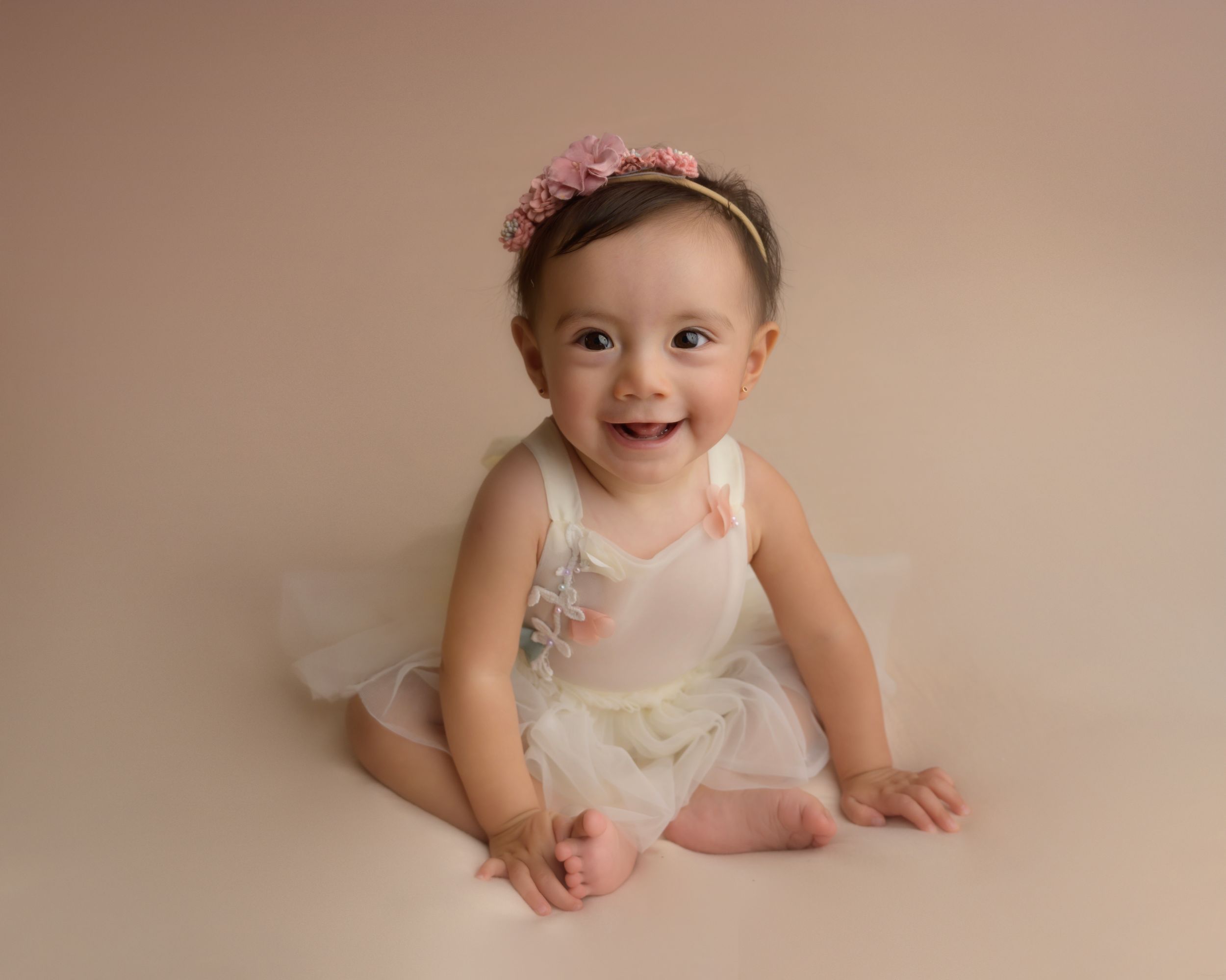 Baby wearing pink floral headband and white dress poses on beige backdrop during professional photo session.