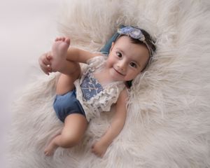 Baby wearing blue bow headband and lace romper lies on white fur blanket during portrait session.