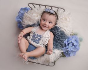 Baby in lace outfit poses on textured blanket with blue hydrangeas during professional photo shoot.