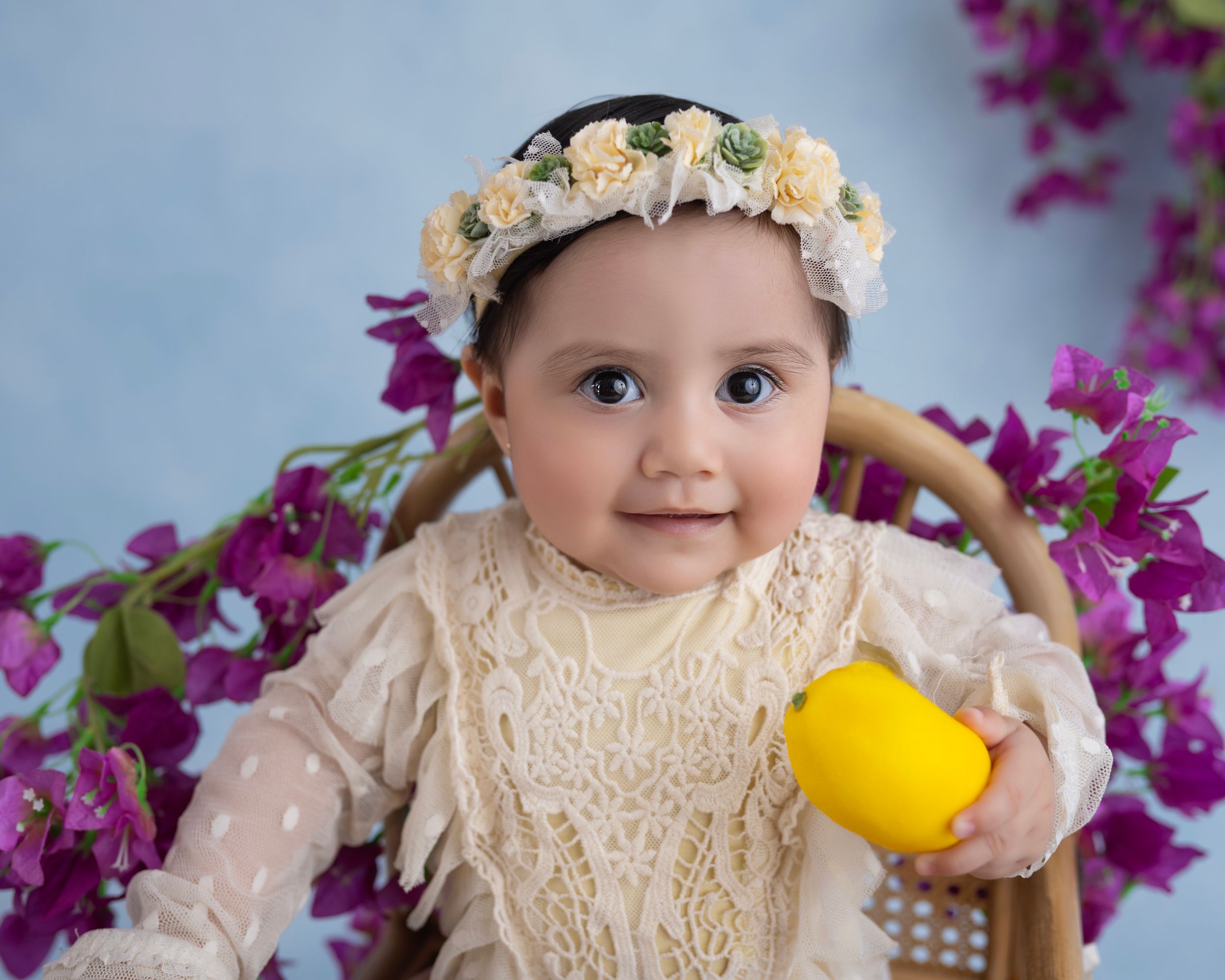 A small child wearing a cream lace dress and floral crown holds a yellow toy against a backdrop of purple flowers.