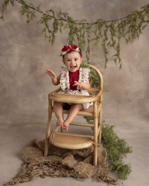 Baby sitting in wooden high chair under draped greenery with burlap floor styling.