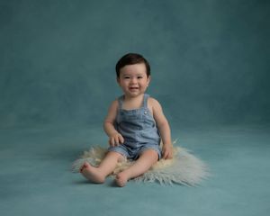 Baby in denim overalls sits on furry rug against teal studio backdrop.