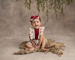 Baby in floral dress with red bow sitting on burlap under hanging greenery in studio setting.
