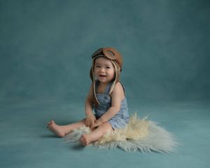 Baby in vintage aviator cap and overalls poses on fur against teal background.