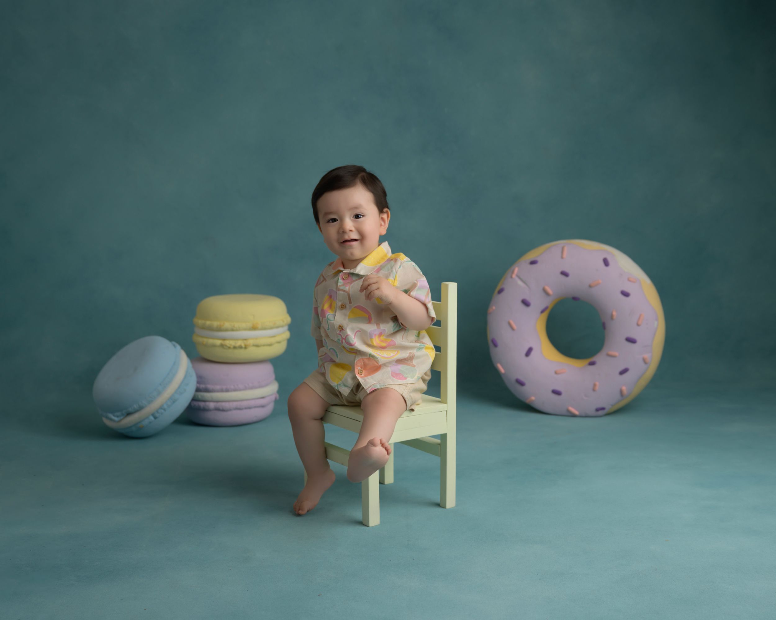 Baby seated on chair with decorative macaron and donut props against teal backdrop.