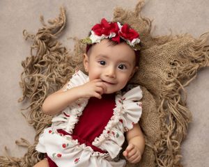 Baby in white and red dress with floral crown lying on textured burlap backdrop.