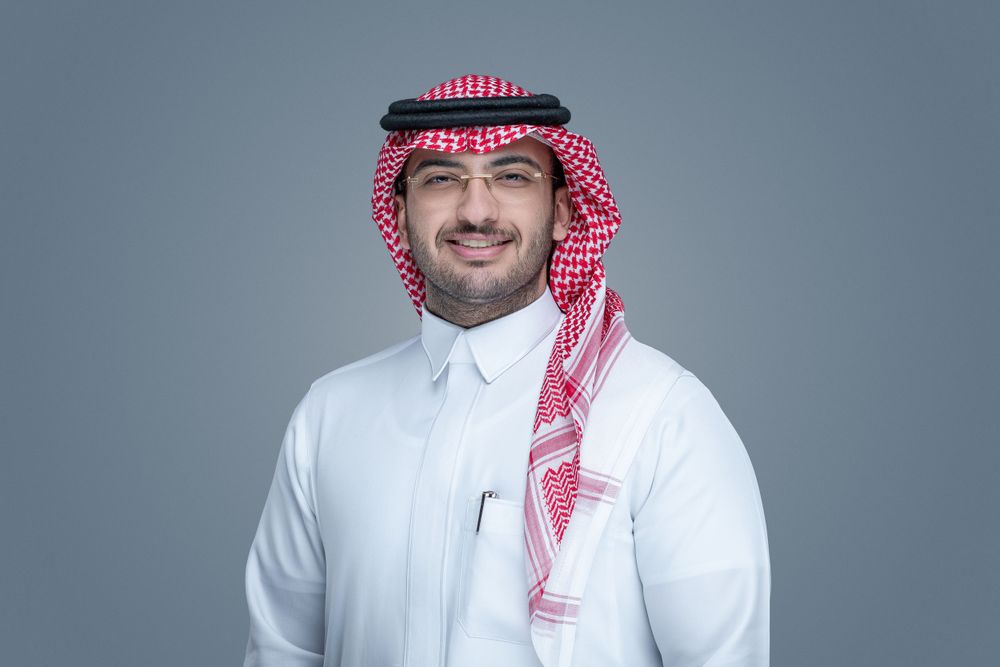 A person wearing traditional Middle Eastern clothing with white thobe and red and white checkered keffiyeh against grey background.
