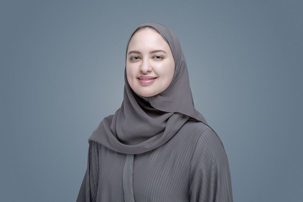 Woman in gray hijab and ribbed top smiles against plain gray background.