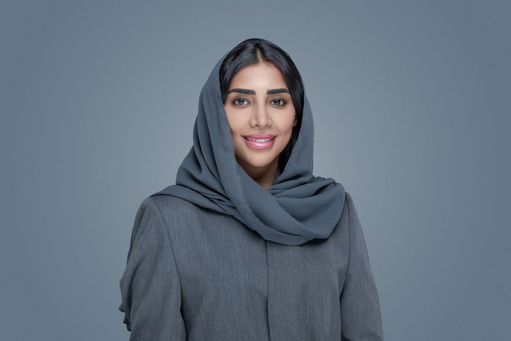 A person wearing a gray hijab smiles warmly against a gray studio backdrop.