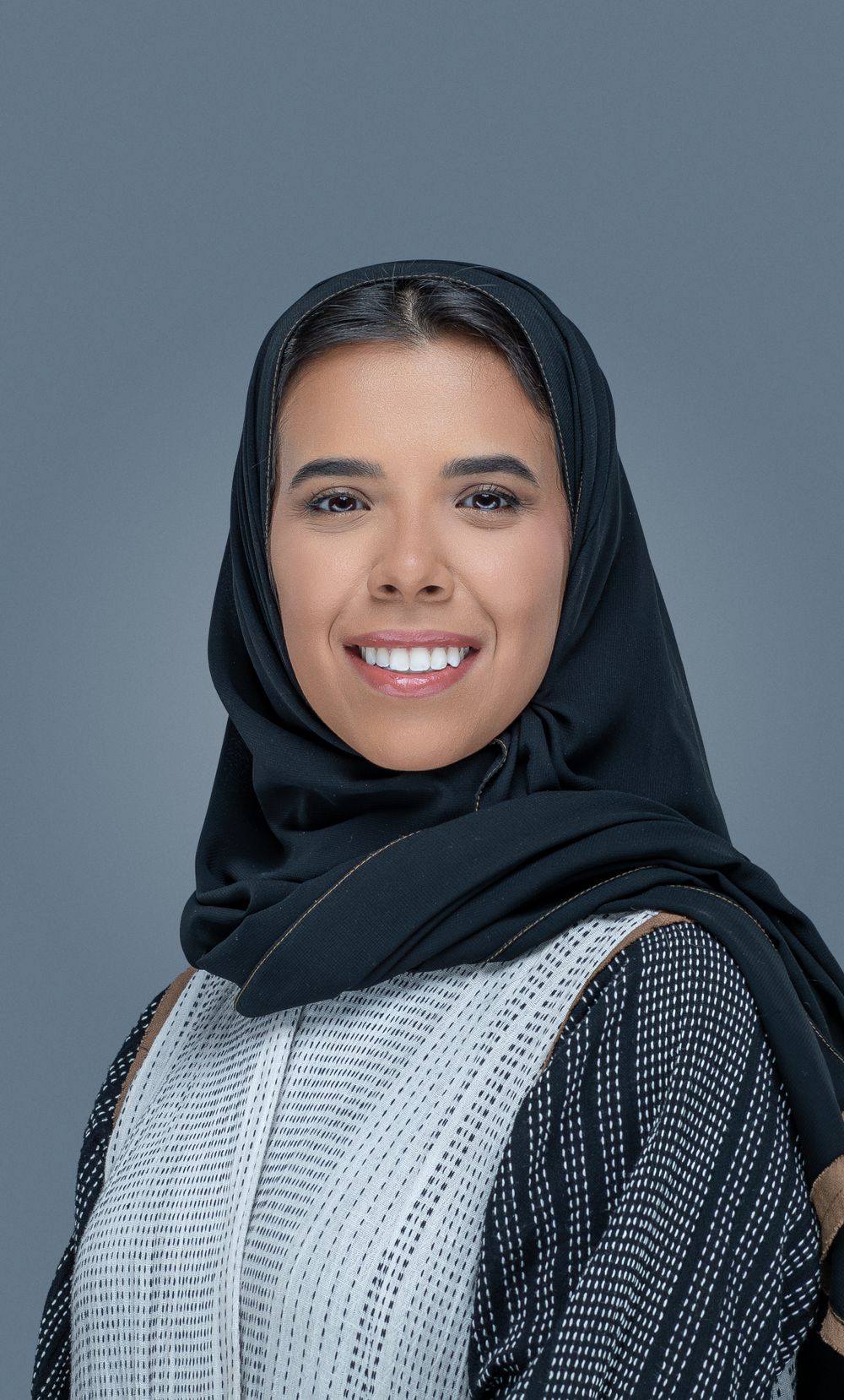 Smiling woman wearing a black hijab and white patterned dress against a grey background.