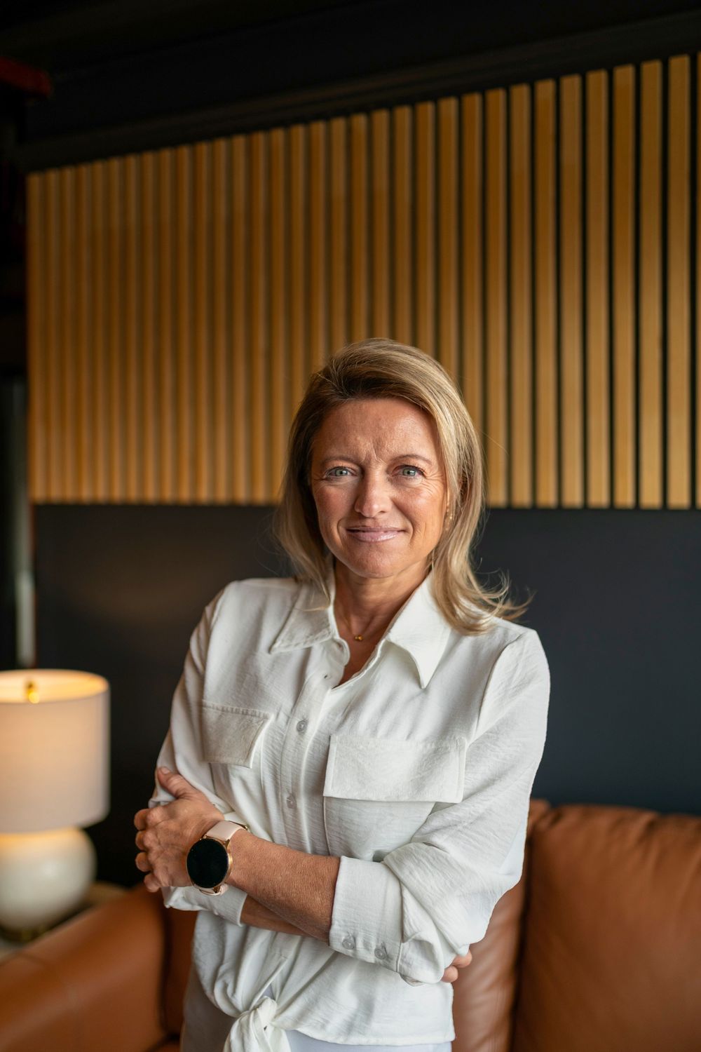 Smiling middle-aged blonde woman in white shirt, arms crossed, standing in modern interior with wood panel wall.