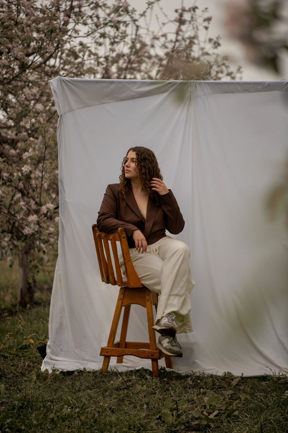Person in brown blazer and white pants poses artistically on wooden chair against white backdrop in outdoor spring setting.