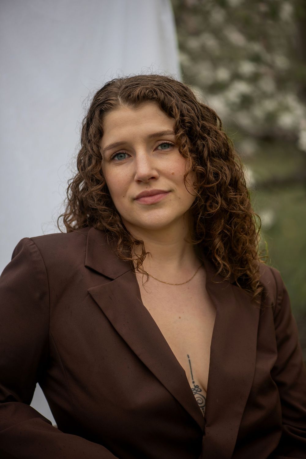 Professional portrait of someone with curly hair wearing a brown blazer and delicate necklace against a blurred outdoor backdrop.