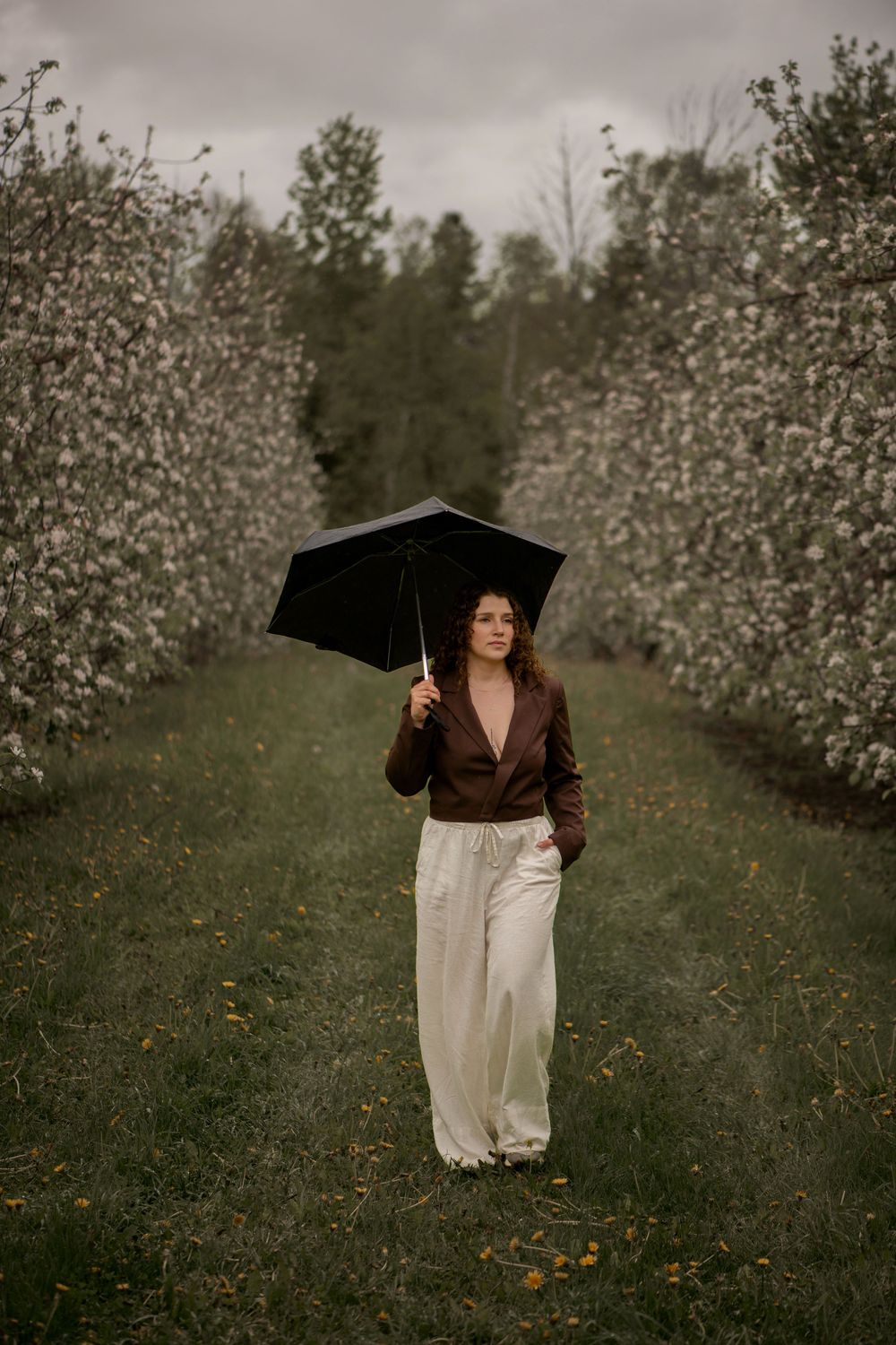 Figure holding black umbrella stands in orchard pathway wearing brown jacket and white pants on moody autumn day.