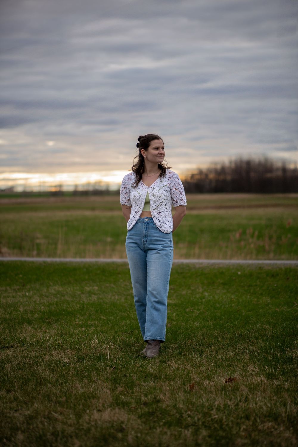 A person stands in a grassy field at dusk wearing a white floral top and light blue jeans.