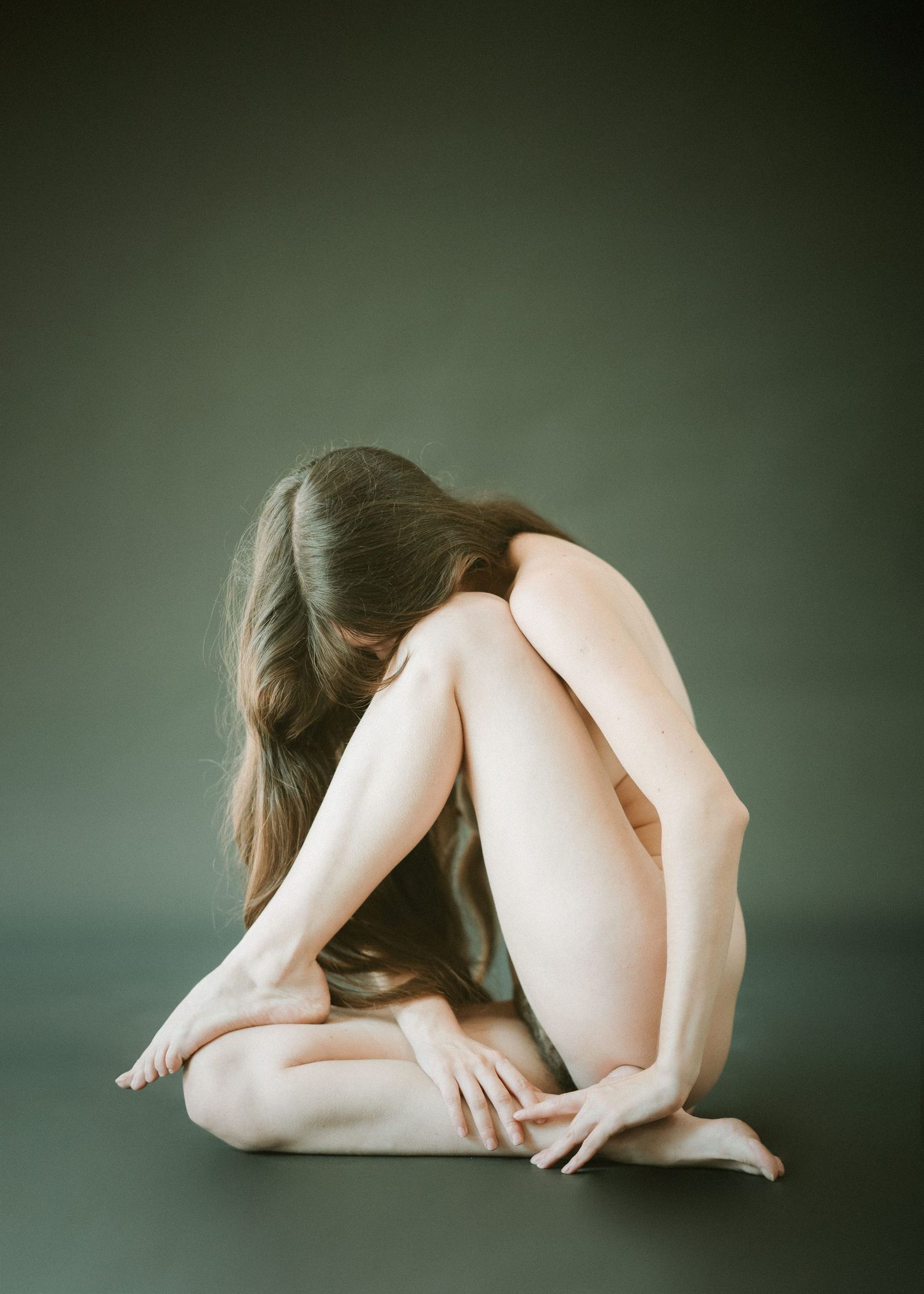 Graceful minimalist artistic nude figure curled in contemplative pose against dark green background.