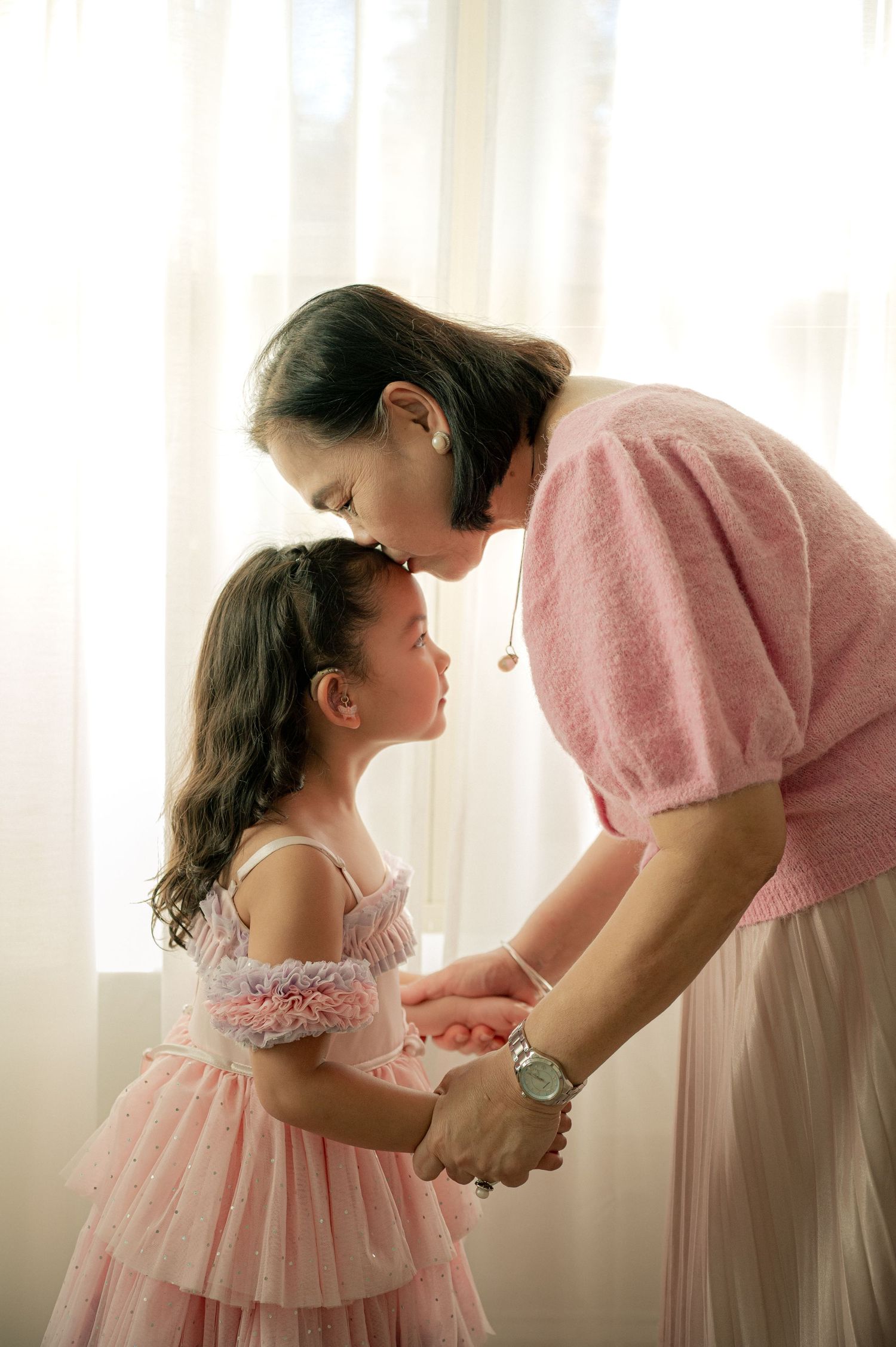 Sweet moment between two people in pink dresses holding hands in front of sheer curtains.