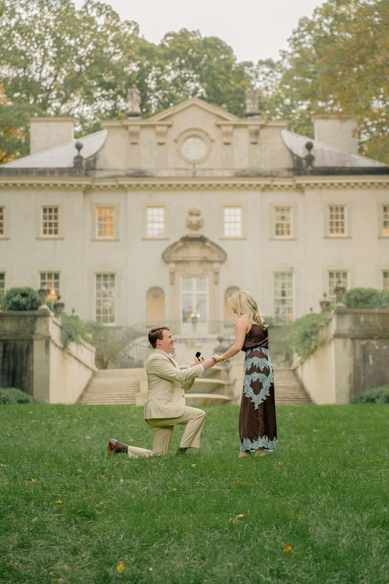 Playful couple poses on grass lawn in front of elegant white mansion building.