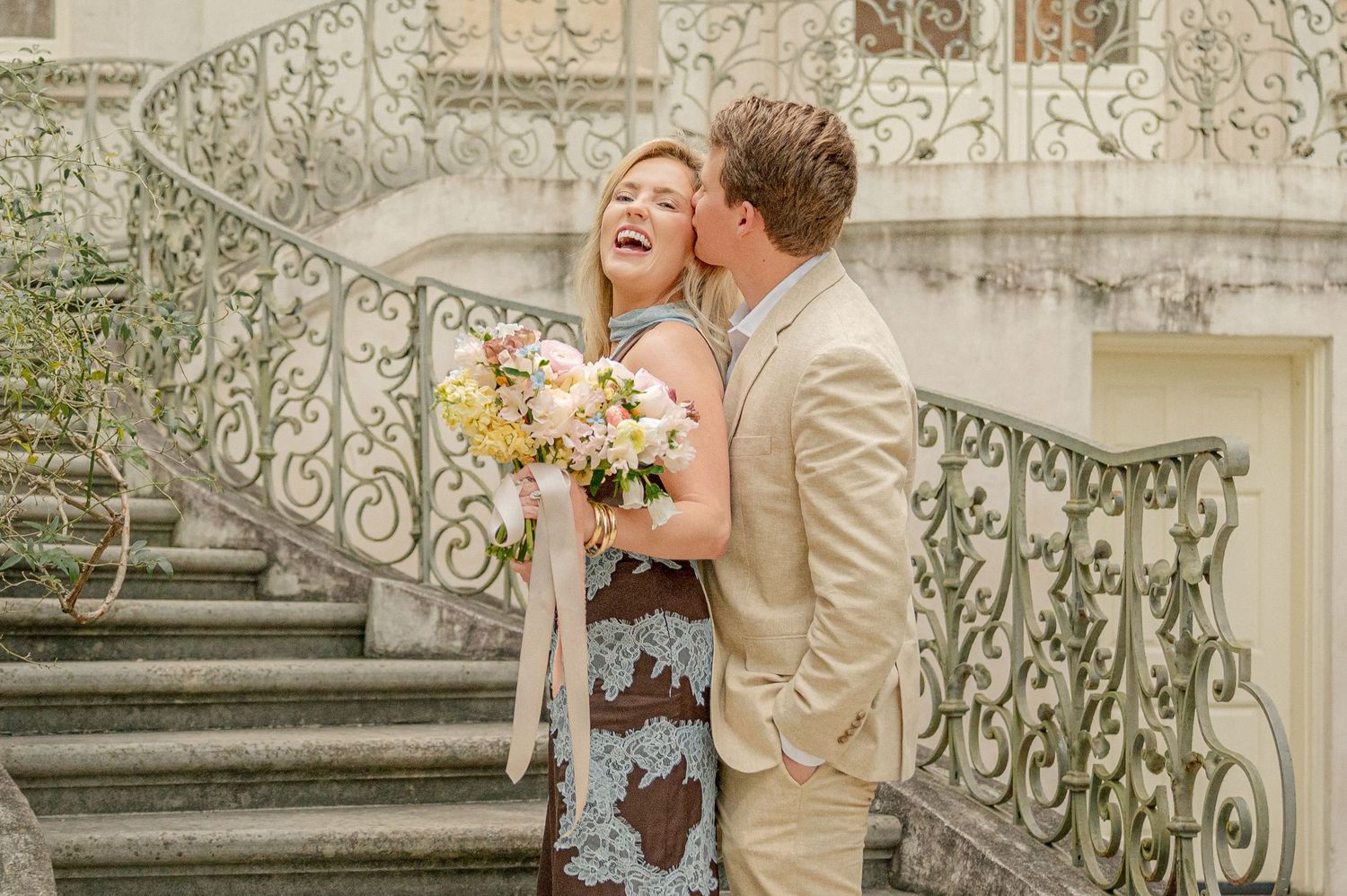 Couple posing on ornate stone staircase with decorative wrought iron railing.
