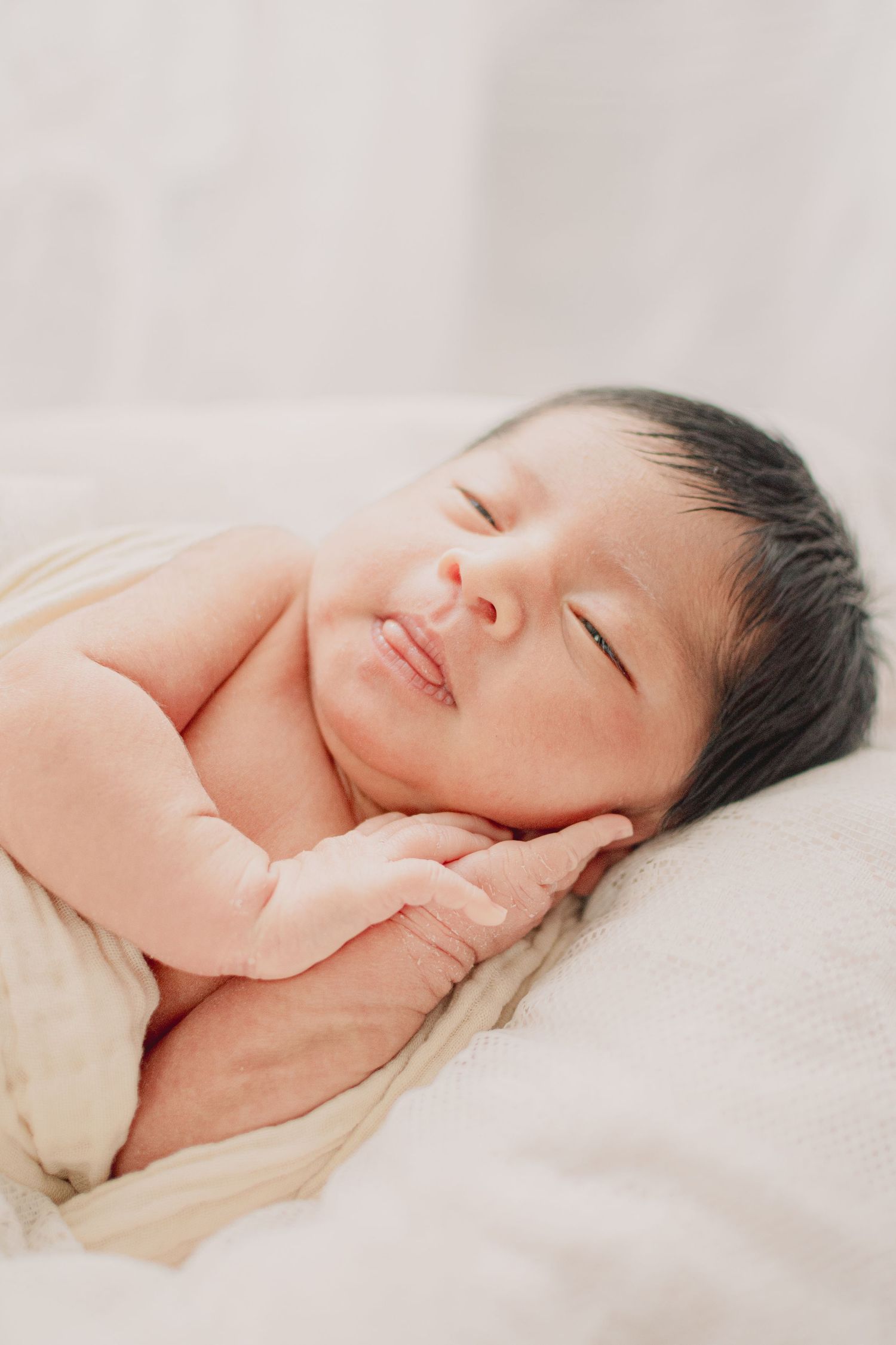 Peaceful sleeping newborn with hands clasped under chin on soft white bedding.